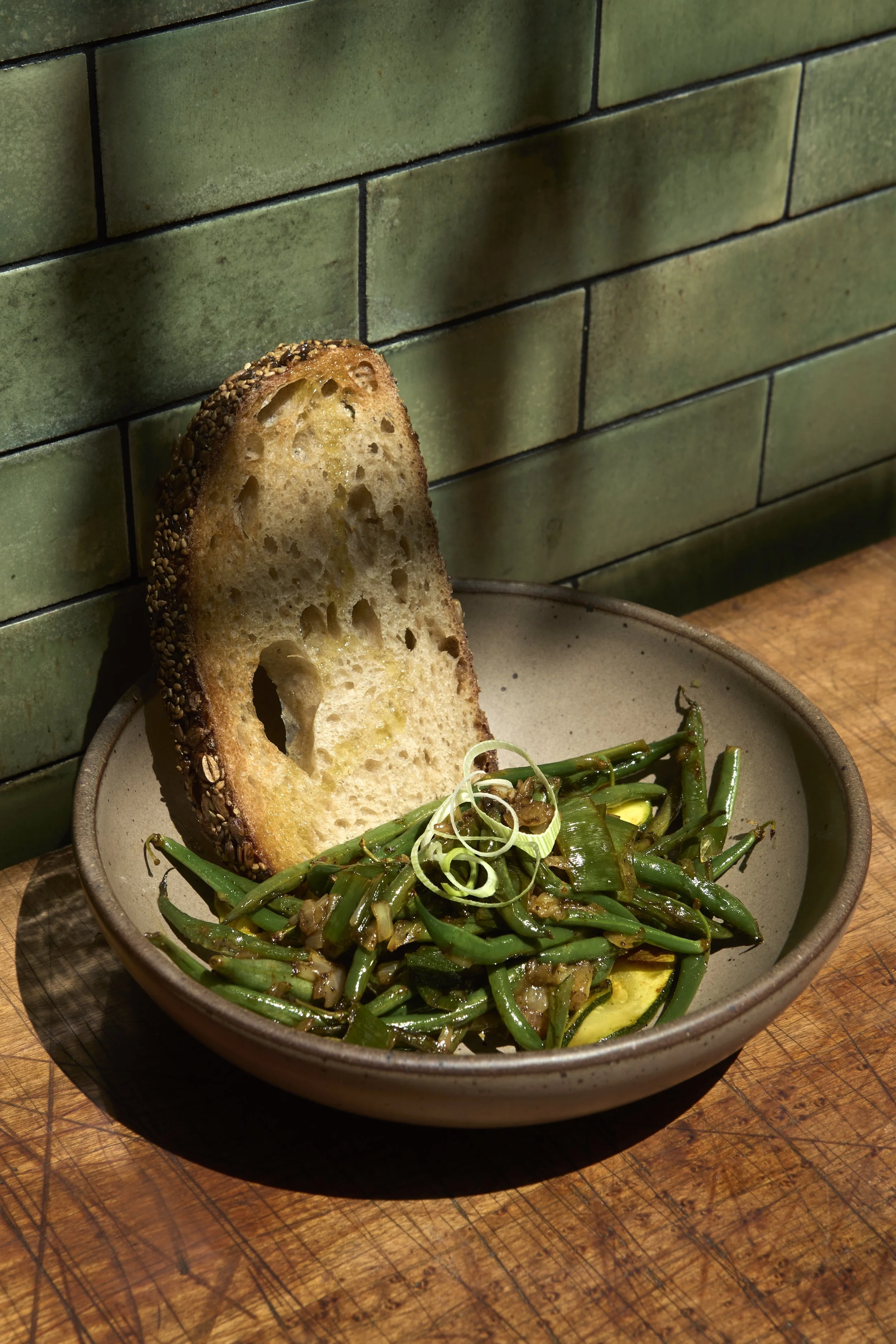 A bowl of sautéed green beans garnished with sliced green onions, served with a slice of toasted bread with sesame seeds, is placed on a wooden surface against a green tiled wall.
Food Photography Los Angeles.