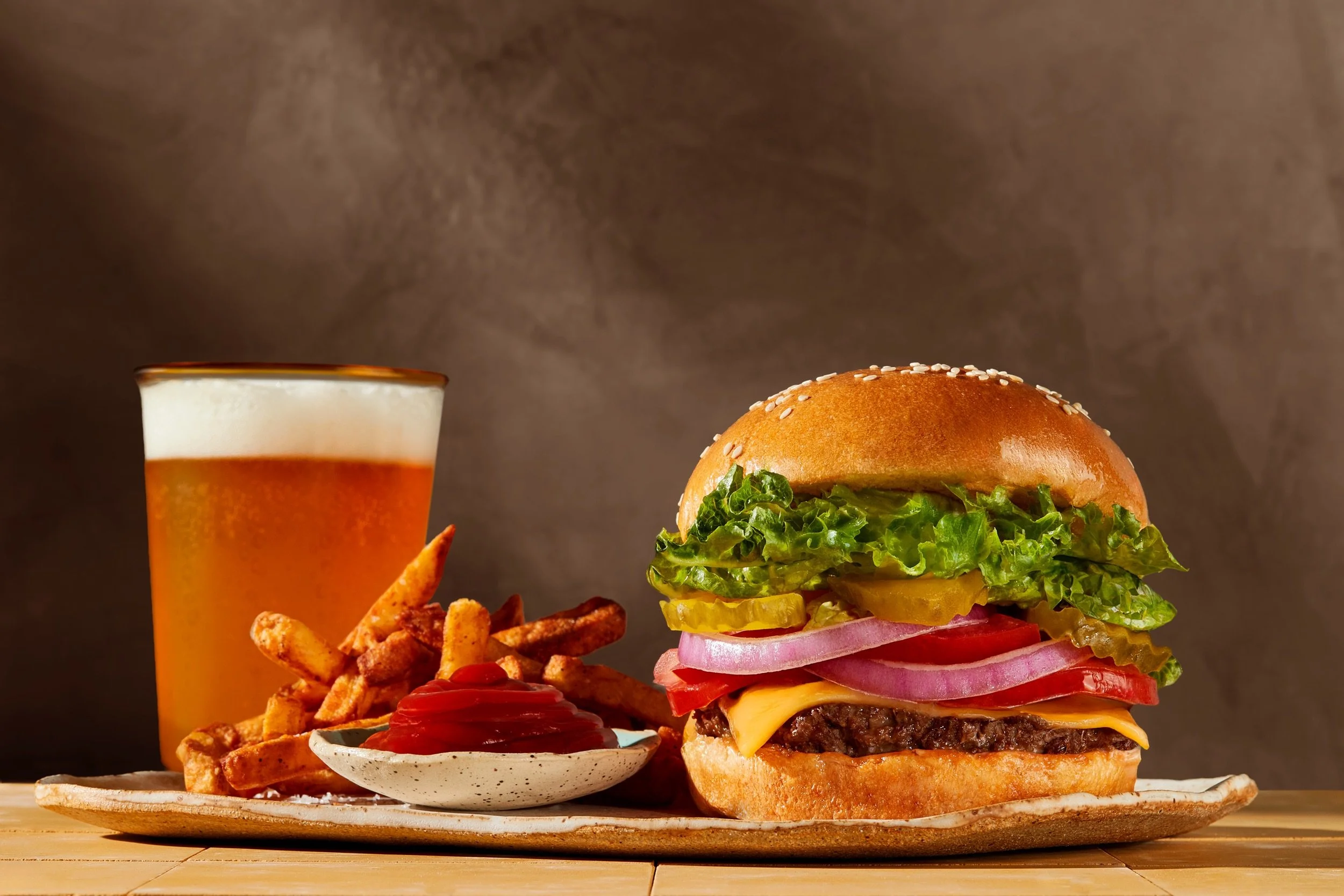 A plate with French fries, ketchup, a cheeseburger with lettuce, tomato, pickles, onions, cheese, and beef patty, a glass of beer in the background. Food Photography Los Angeles.