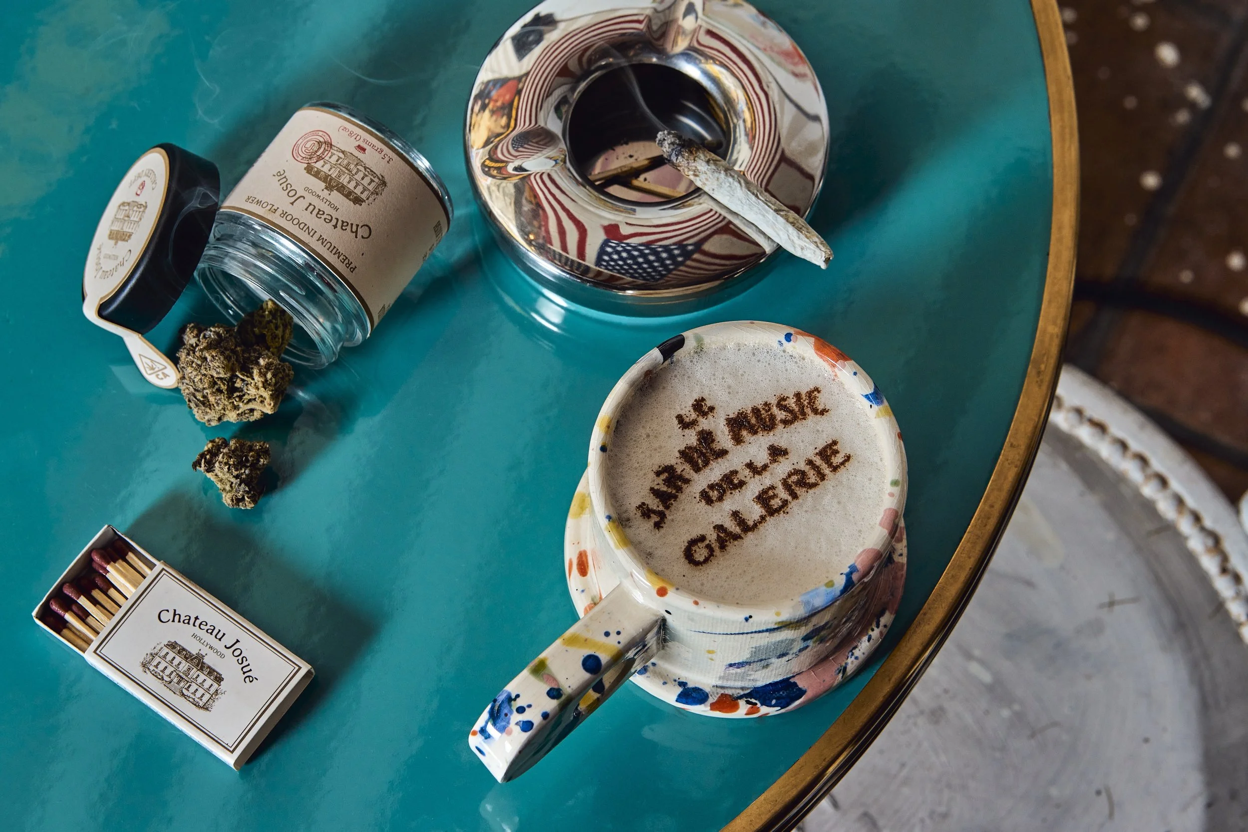 A turquoise table with a colorful mug of foamy coffee, a matching saucer, a glass ashtray with a lit cigarette, a jar of cannabis, and a pack of matches. Los Angeles beverage and food photography.