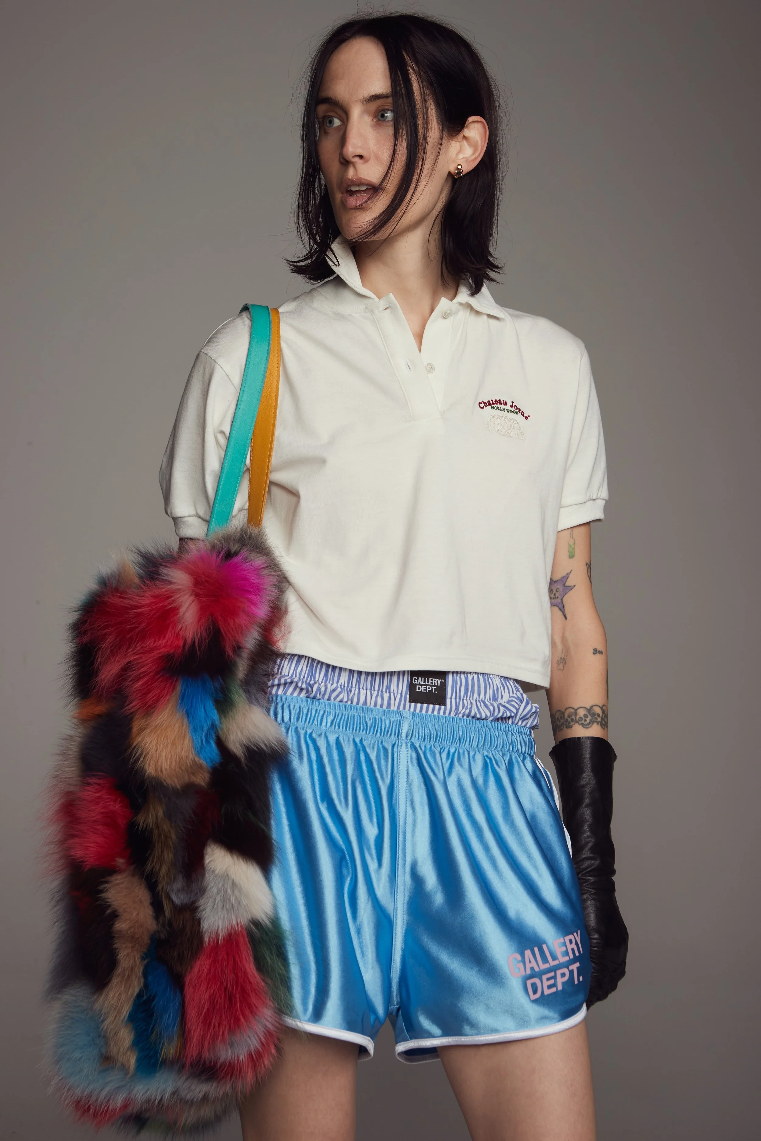 A young woman with dark hair and tattoos wearing a white Gallery Department polo shirt, bright blue shorts, black gloves, and carrying a multi-colored furry bag against a gray background.  Los Angeles On-Model fashion photography.