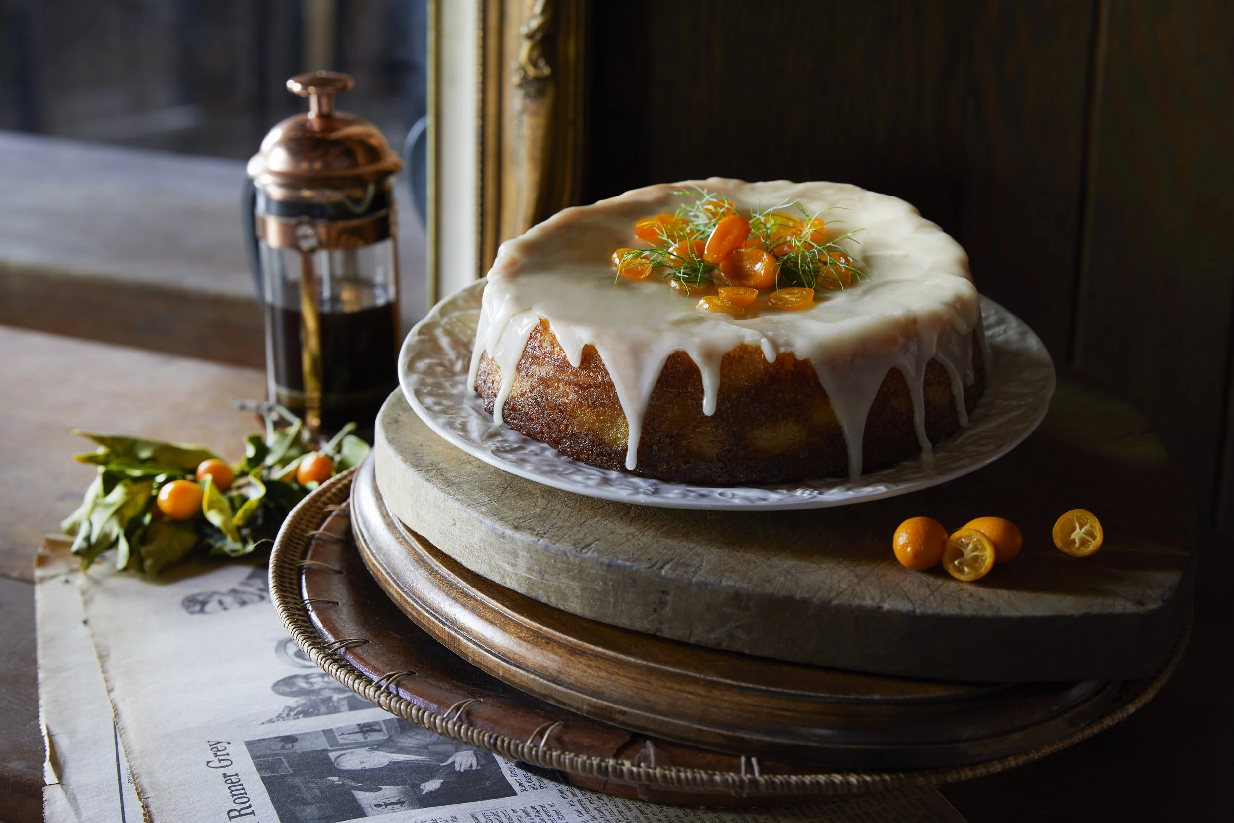 Coffee cake with white icing and orange berries on top, on a white plate, placed on a wooden stand with a wooden table, a jar of syrup or honey, and an arrangement of orange berries and leaves nearby. Los Angeles food photography.