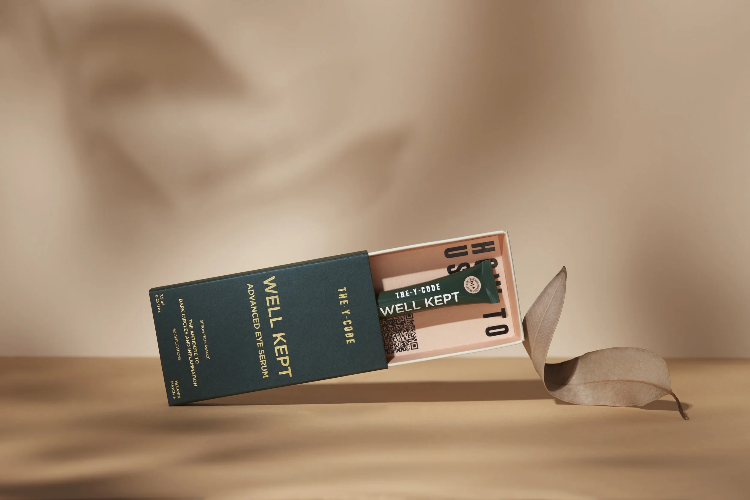 Open box of THE-Y-CODE Well Kept Advanced Eye Serum with a tube inside, resting on a wooden surface, with a dried leaf beside it, in a minimalistic setting. Beaty Product Photography Los Angeles.