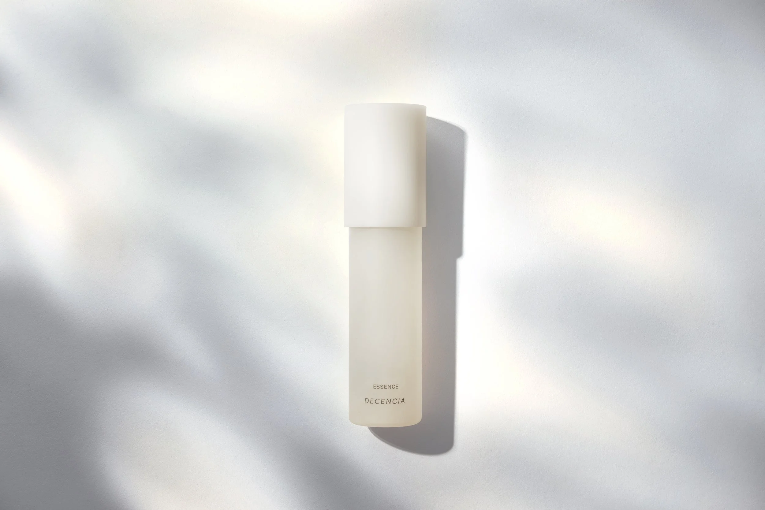 A white cylindrical bottle with a cap, labeled 'ESSENCE' and 'DECENCIA', is placed on a light-colored surface with shadows. Los Angeles beauty product photography.