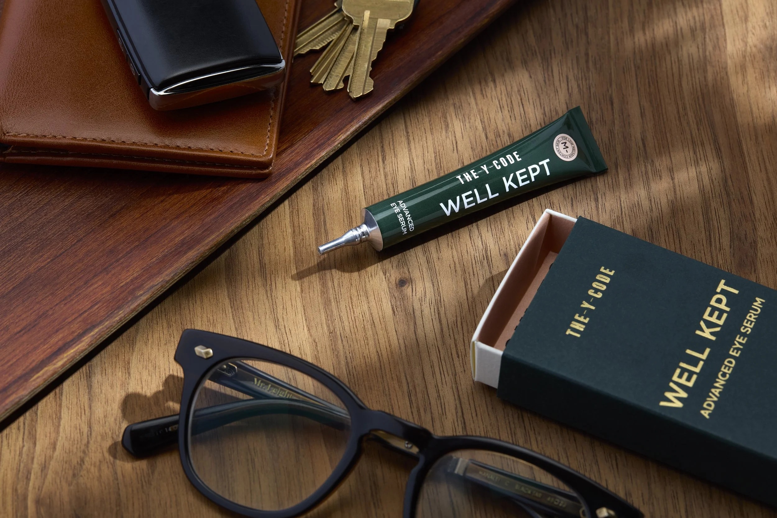 A wooden desk with a box of 'They X Code Well Kept' eye serum, a tube of the serum, black glasses, and a wallet with keys on top. Los Angeles beauty product photography.