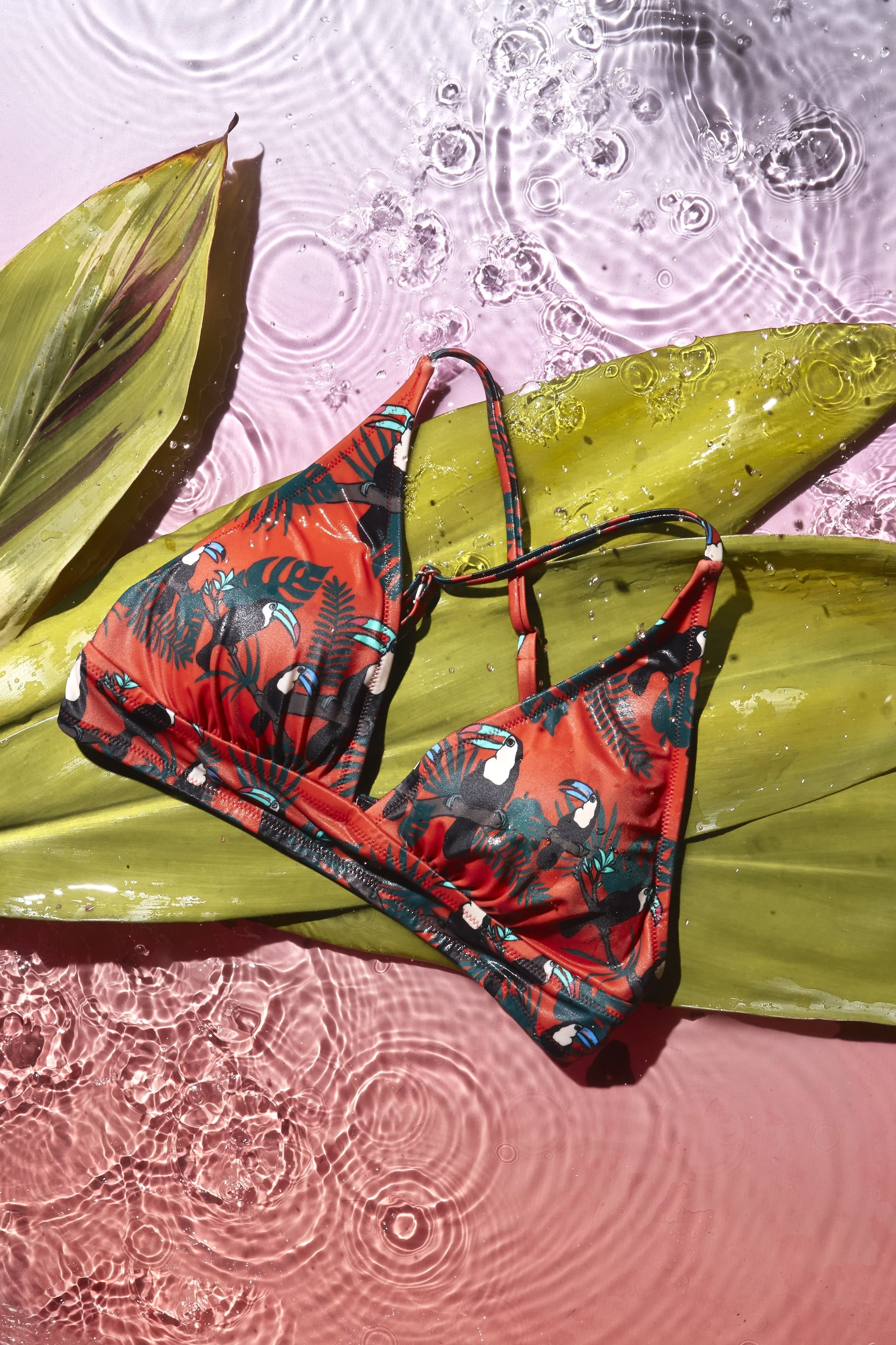 Colorful bikini with toucans and tropical leaves pattern lying on large green leaves and pink water with ripples. Product photography Los Angeles.