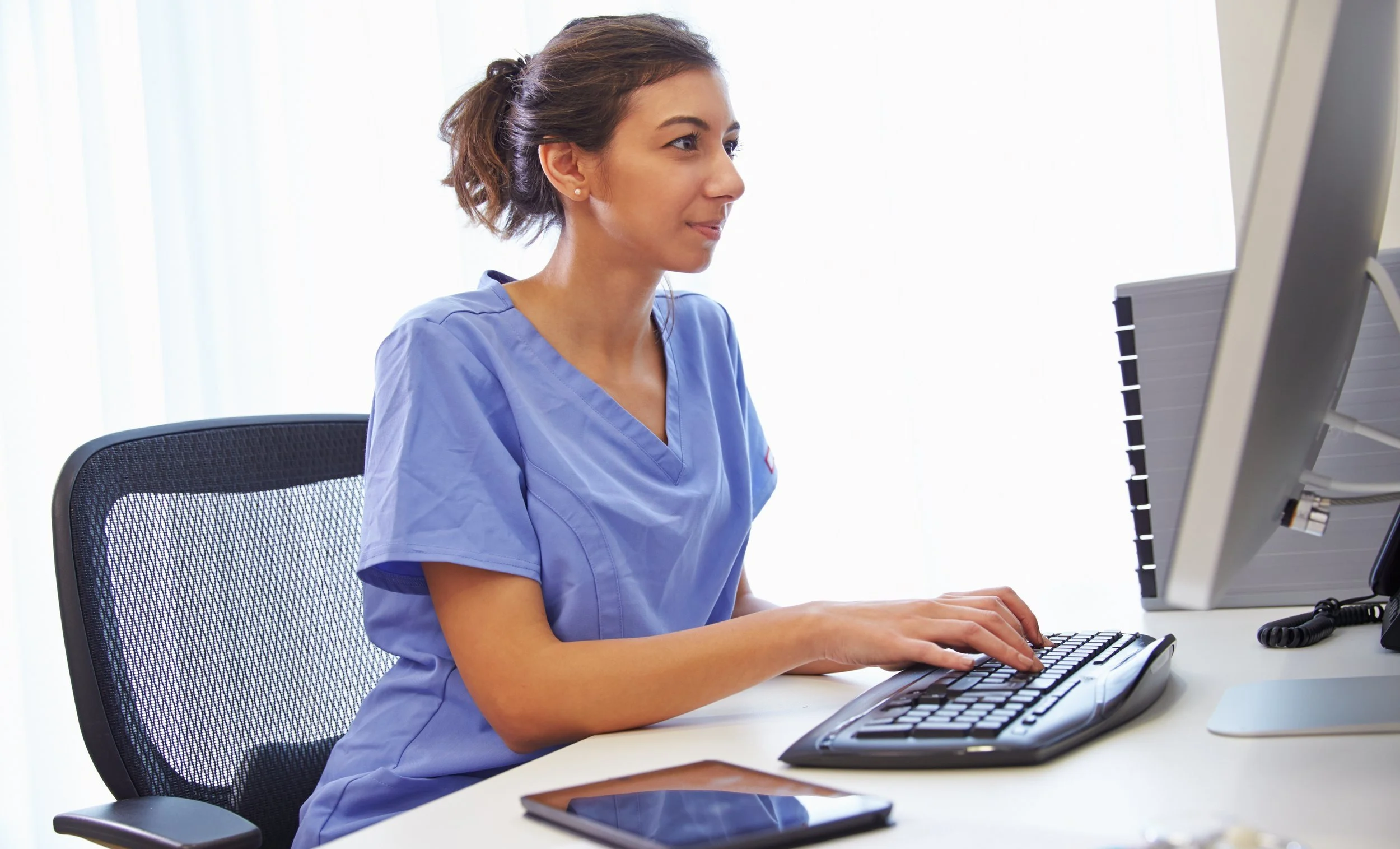 A nurse in blue scrubs working at a computer workstation in an office setting.