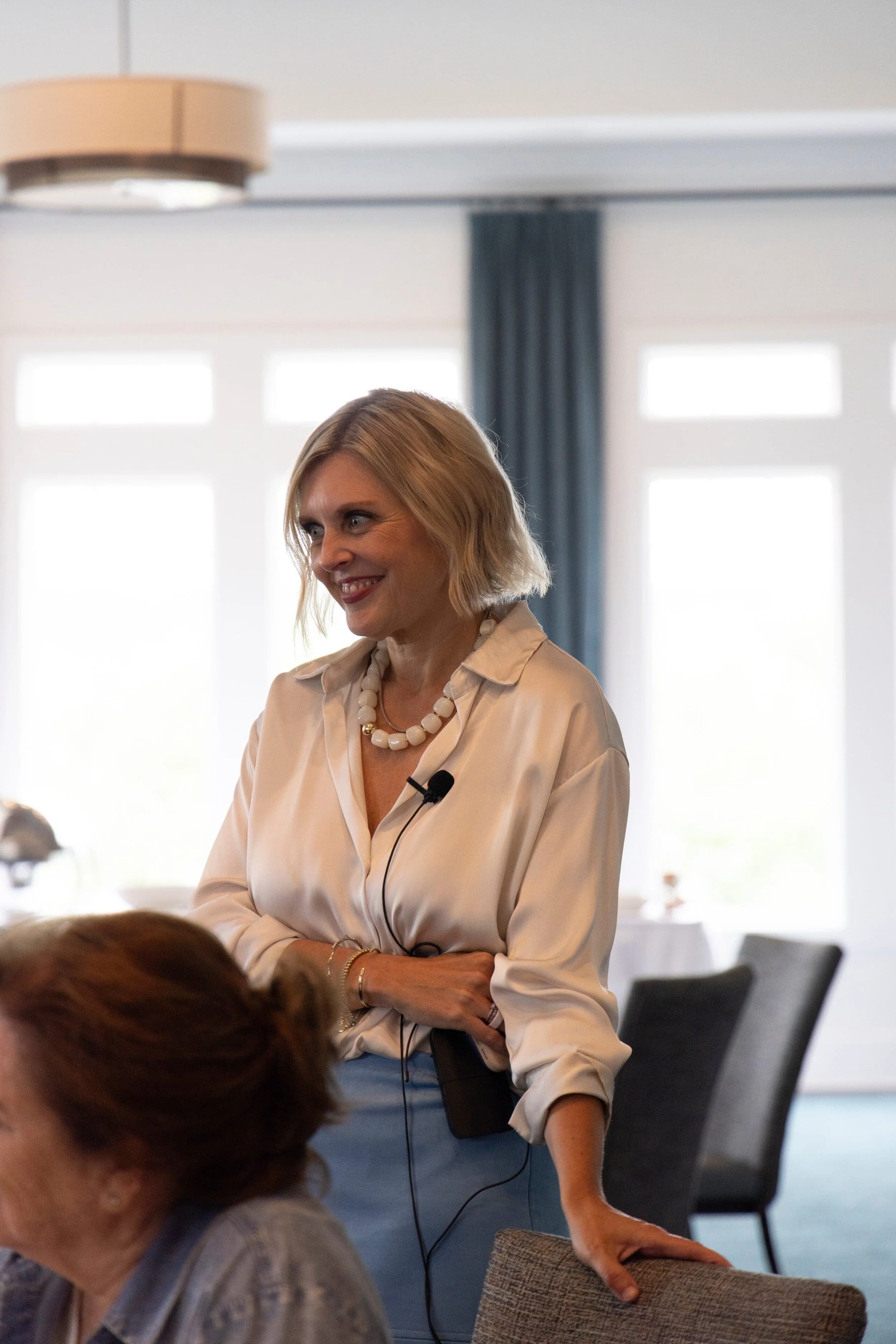 Krista Demcher smiling at a speaking event in a white blouse and blue skirt, wearing a headset microphone