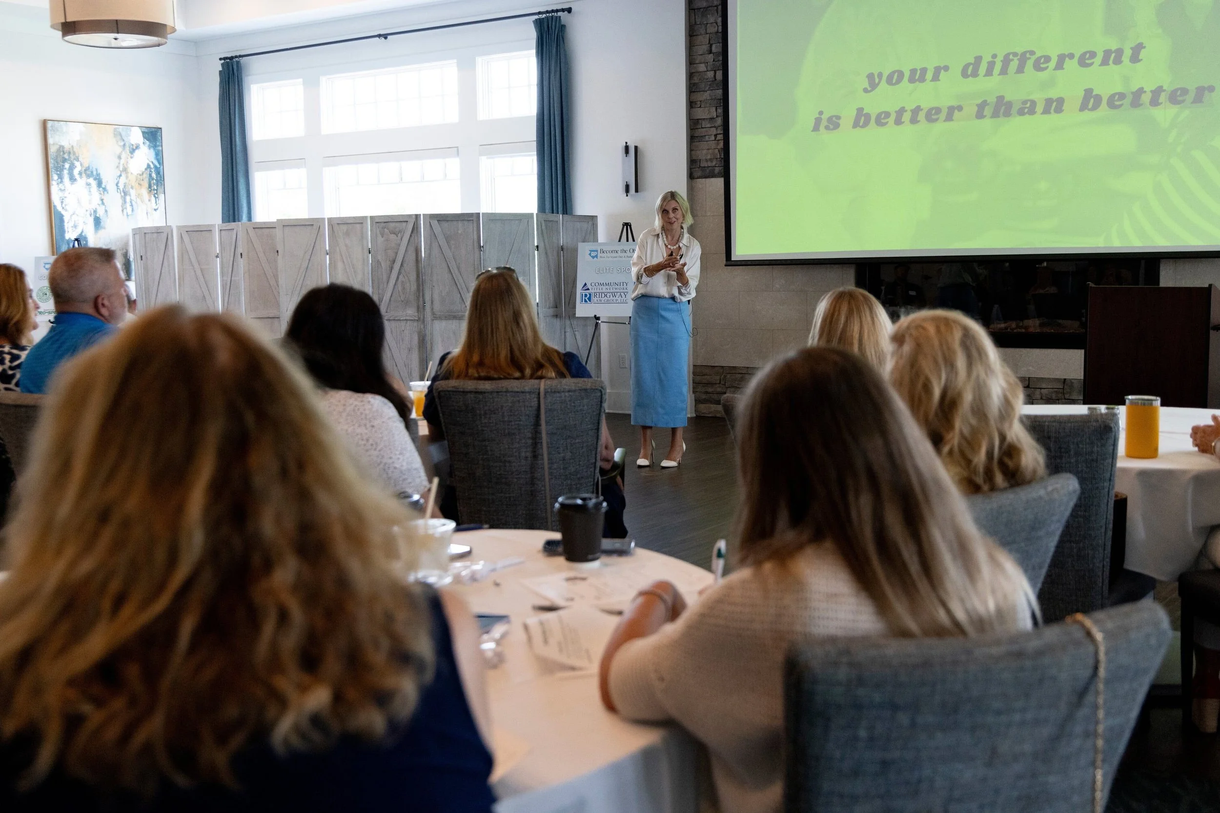 Krista Demcher speaking at the front of a ballroom of seated attendees, with the words your different is better than better behind her