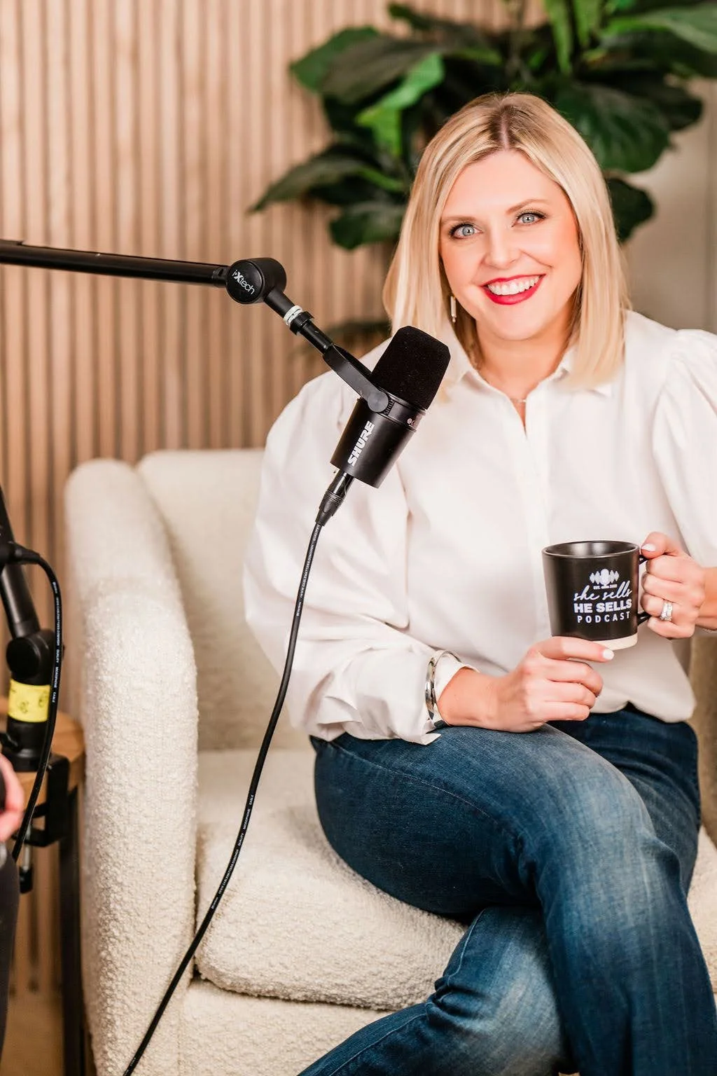 A woman with blonde hair, wearing a white shirt and jeans, sitting on a beige couch, holding a black mug that reads 'the tell HE SELLS PODCAST,' smiling, with a microphone set up in front of her in a podcast recording studio.