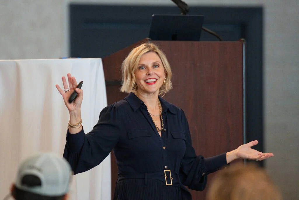 Krista Demcher in a navy dress smiling with one arm outstretched and a presentation clicker in her hand while speaking at a podium