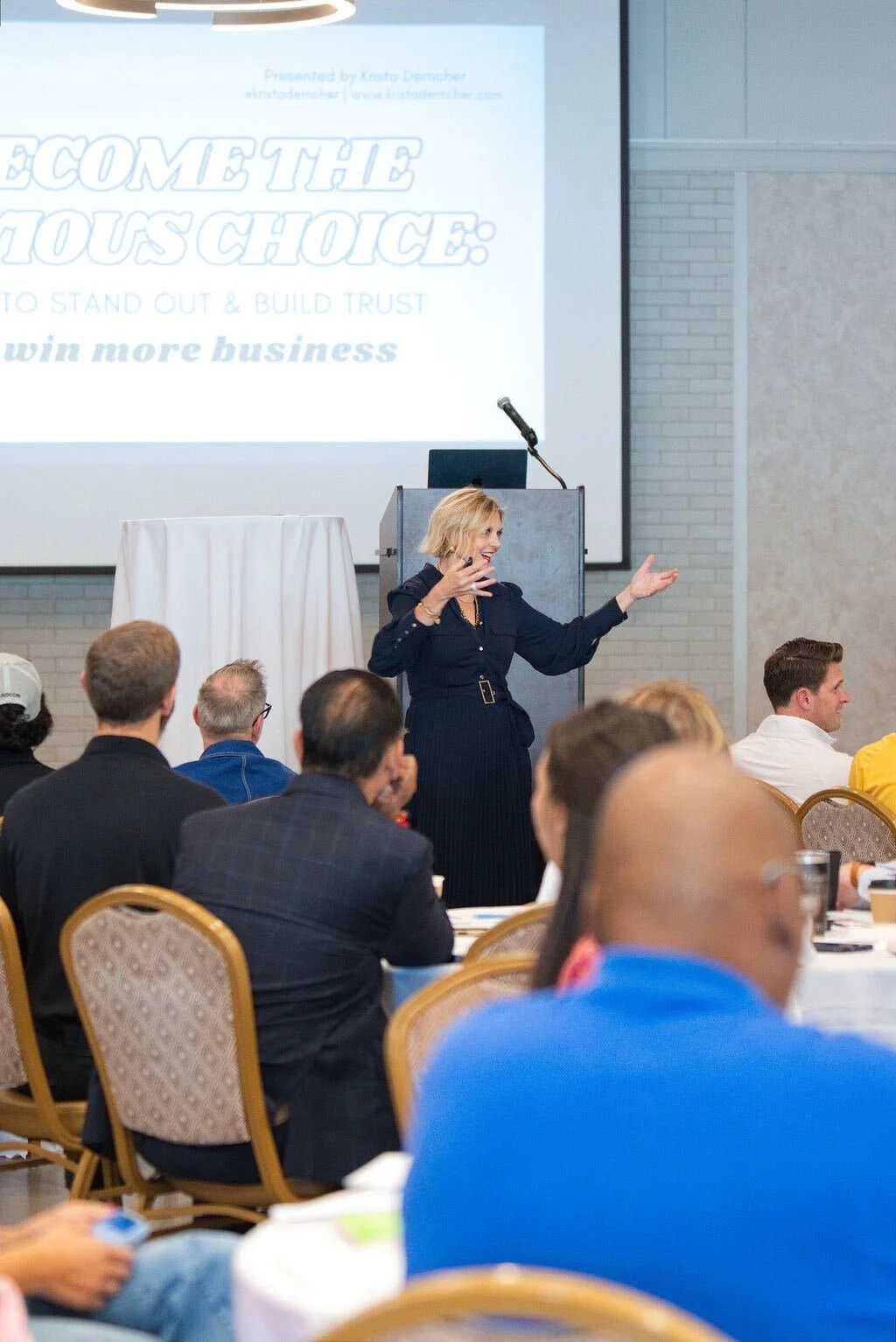 Krista Demcher speaking with open arms at the front of a ballroom of seated attendees, with a slide titled Become the Obvious Choice behind her