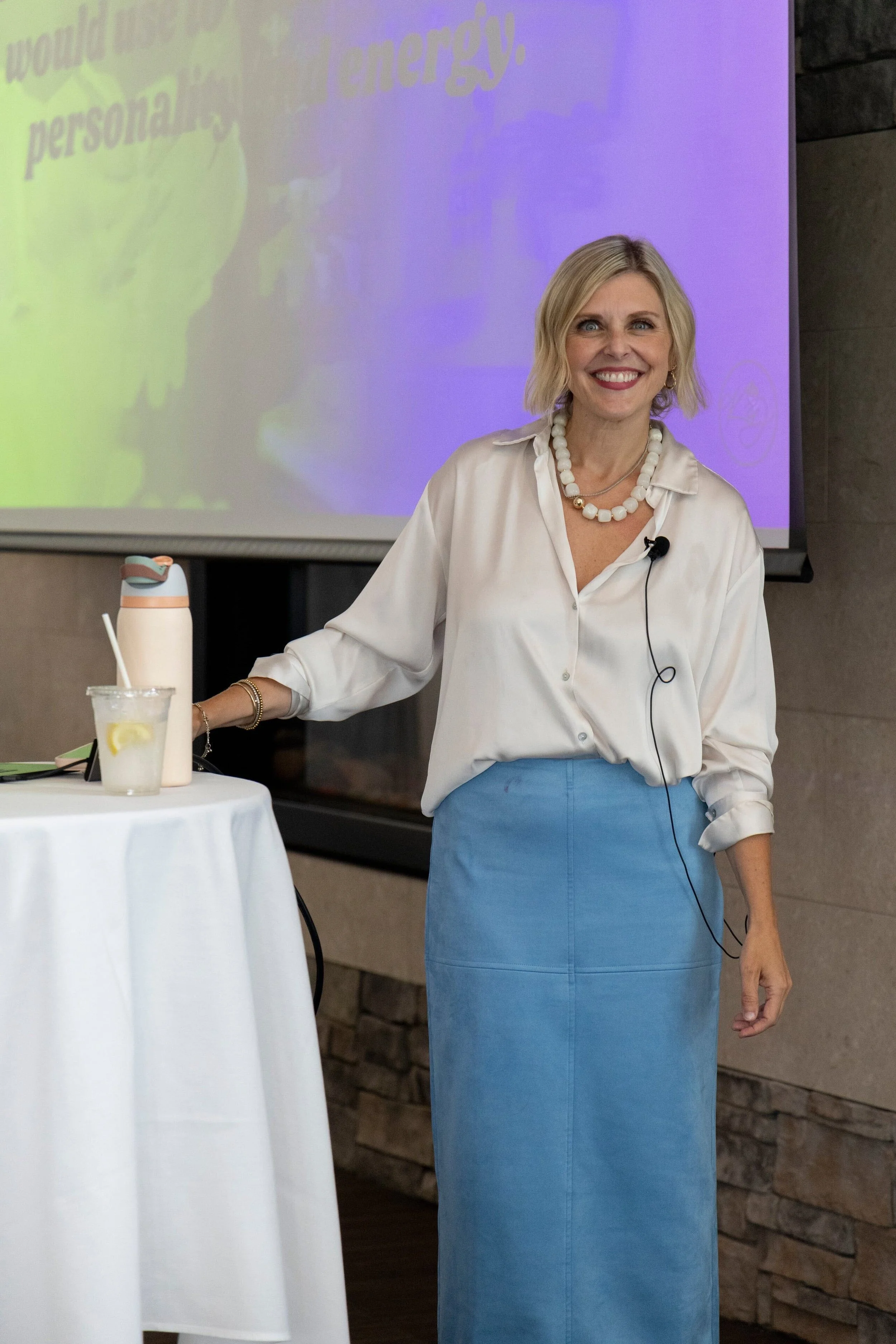 Krista Demcher smiling at a speaking event in a white blouse and blue skirt, wearing a headset microphone