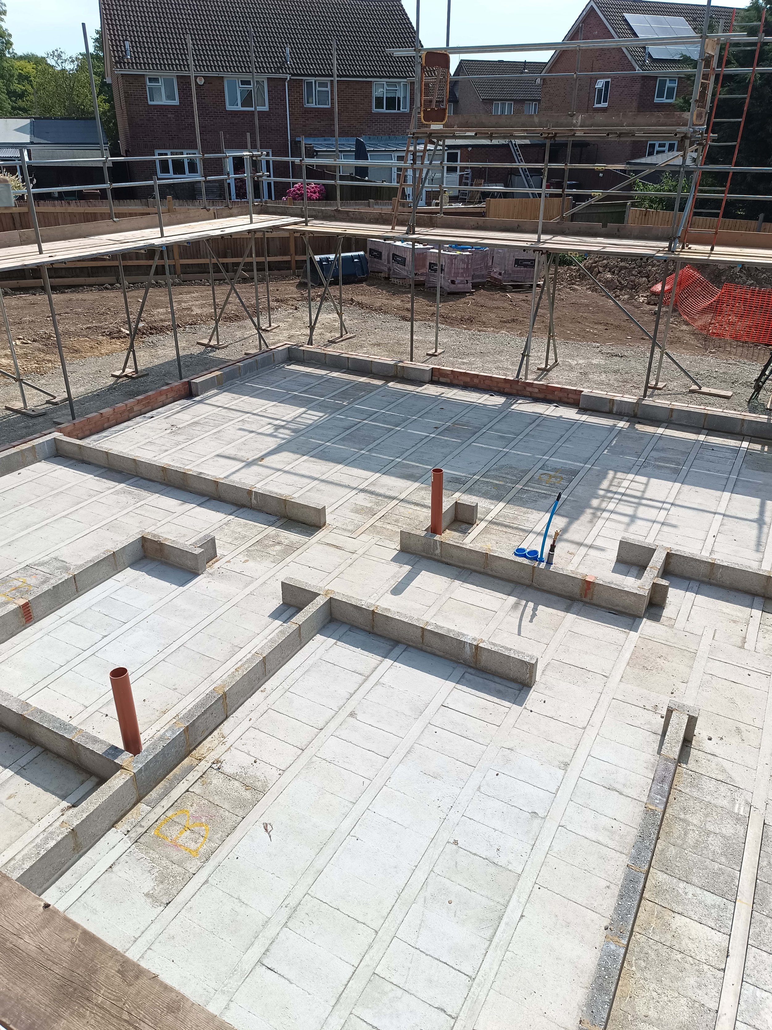 Construction site with foundation work, scaffolding, residential buildings in background.