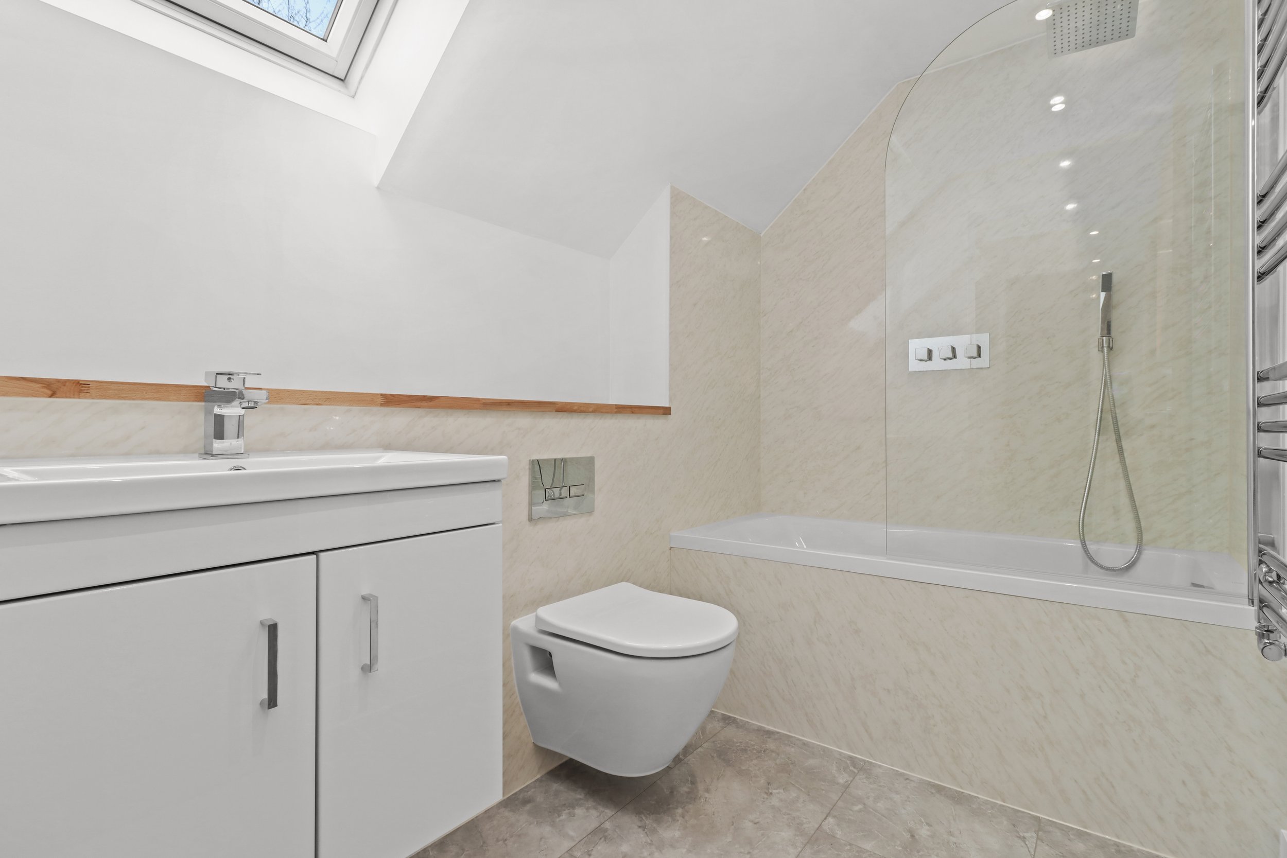 Modern bathroom with white fixtures, including a sink cabinet, wall-mounted toilet, and a bathtub with a glass shower screen. The room has beige tiled walls, a skylight, and a towel radiator on the side.