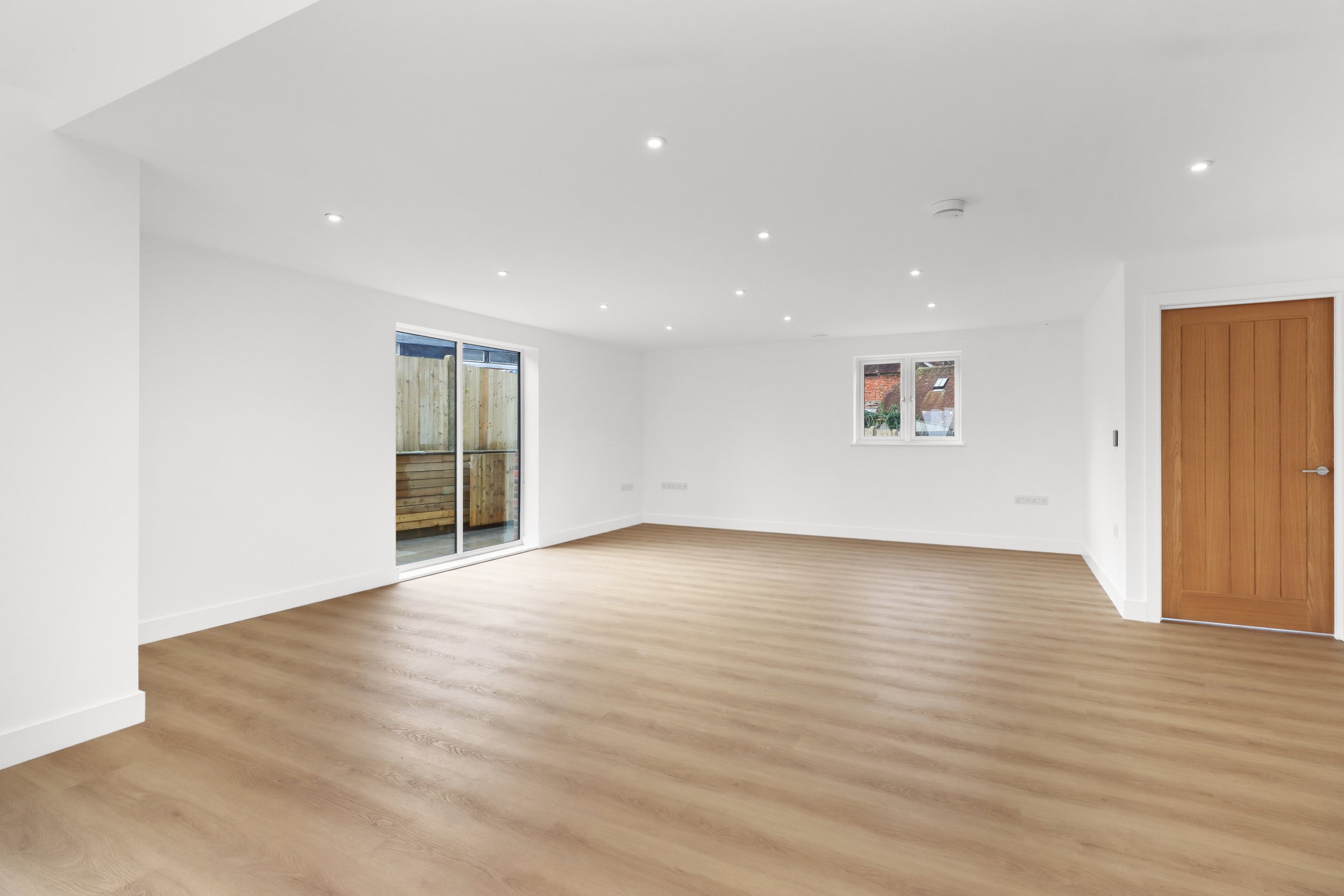 Empty modern living room with wood flooring, white walls, recessed lighting, a wooden door, and a sliding glass door leading to a patio.