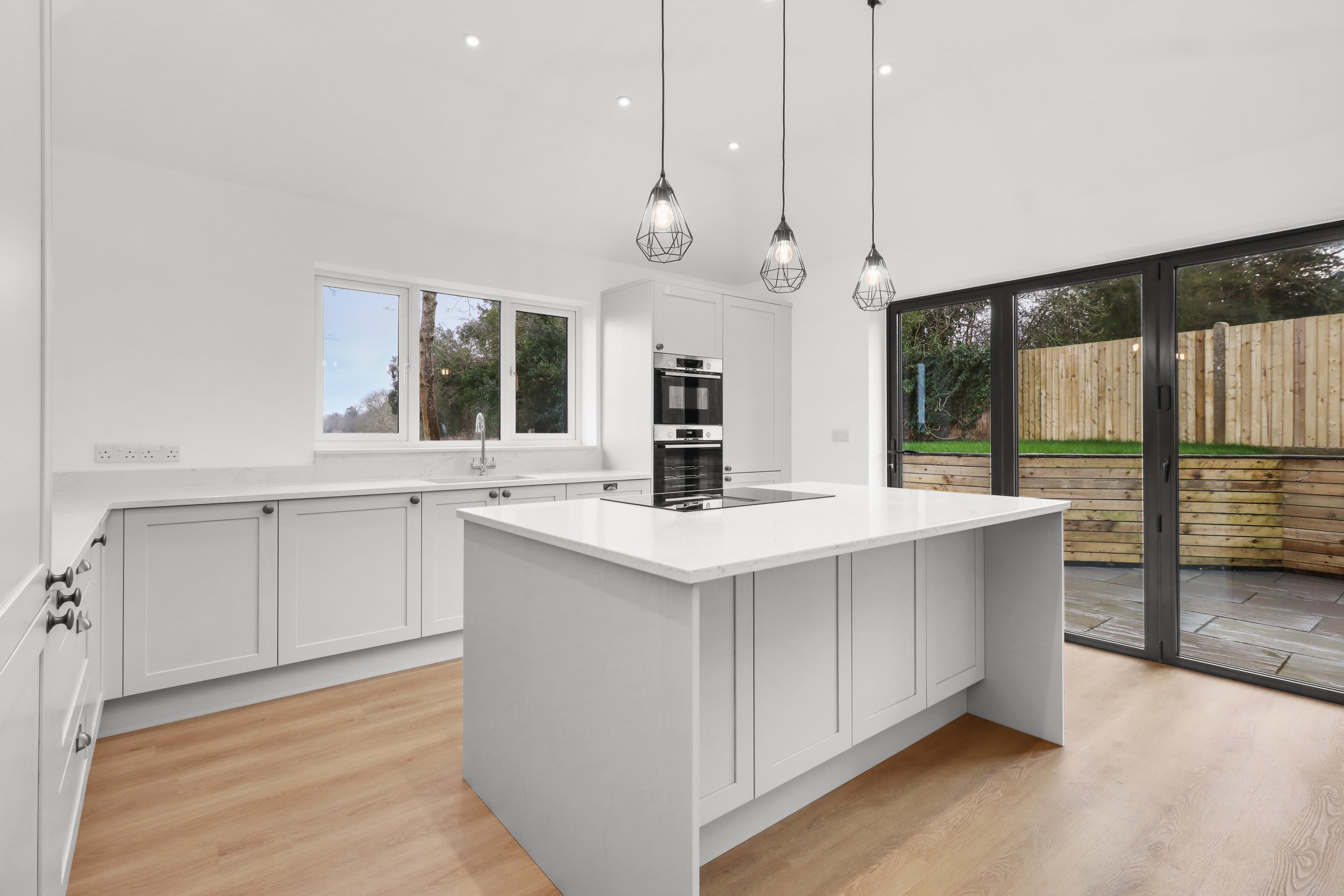 Modern white kitchen with marble countertops, island, built-in oven, pendant lights, wood flooring, and large sliding glass doors.