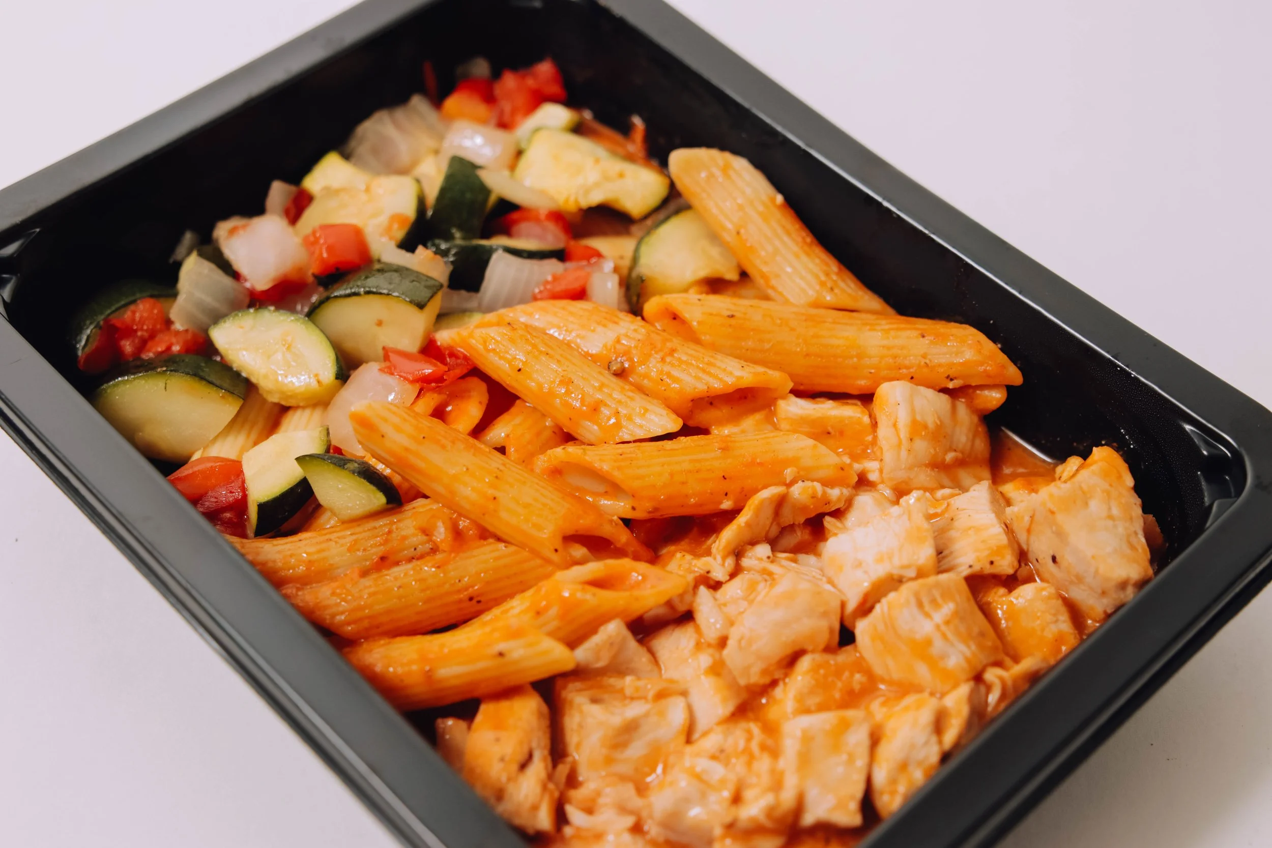 Frozen meal container with cooked penne pasta in red sauce, grilled chicken pieces, and sautéed zucchini, onions, and peppers.