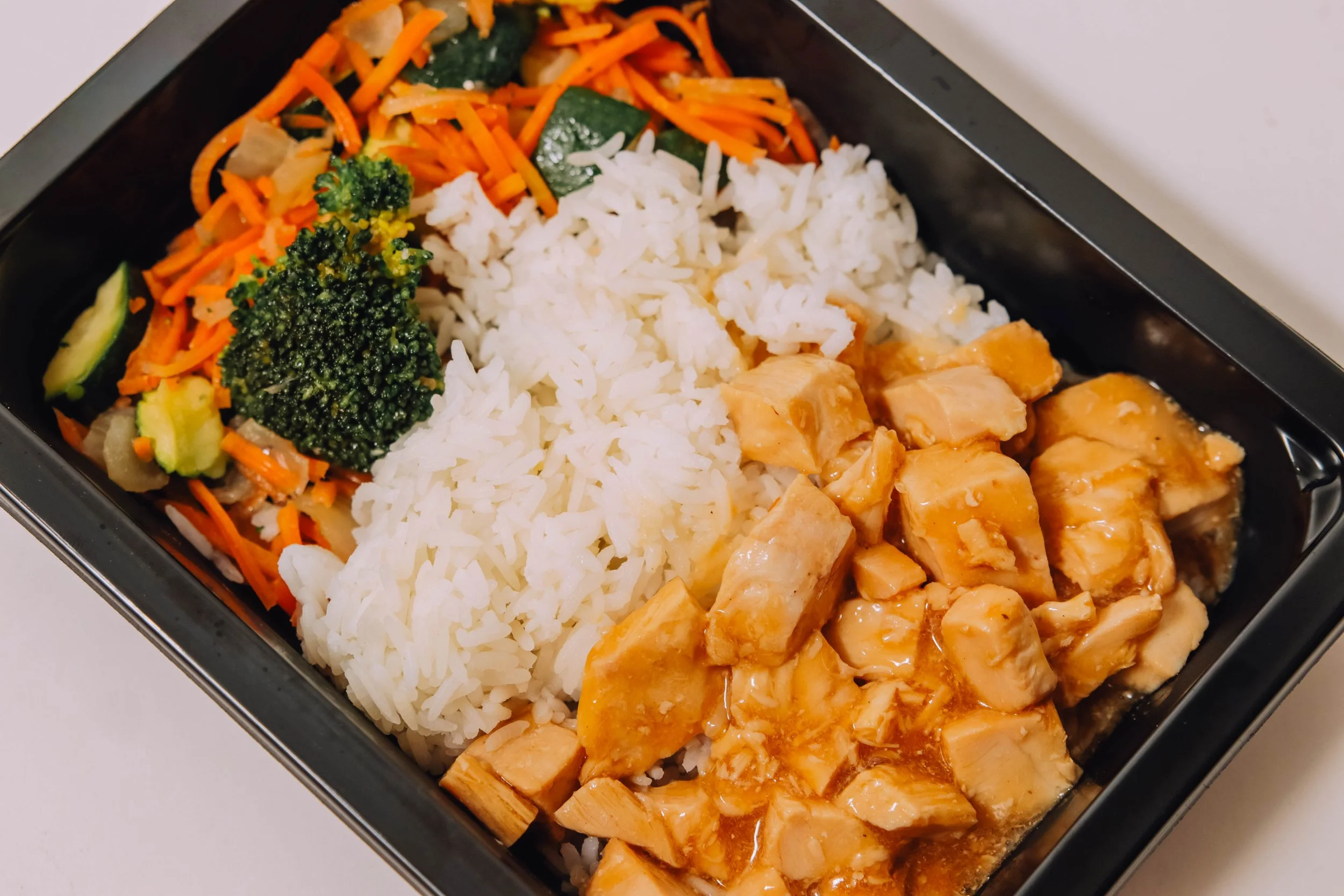A black container with white rice, cubed chicken in brown sauce, mixed vegetables including broccoli, carrots, zucchini, and onions.