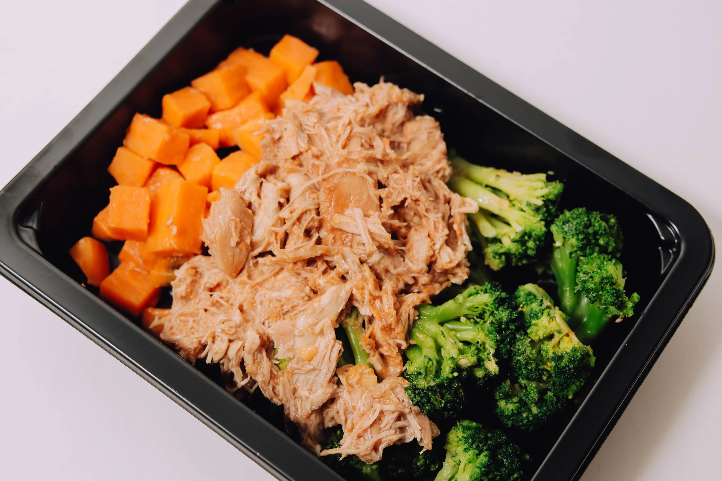 Black takeout container with cooked sweet potatoes, shredded chicken, and broccoli florets.