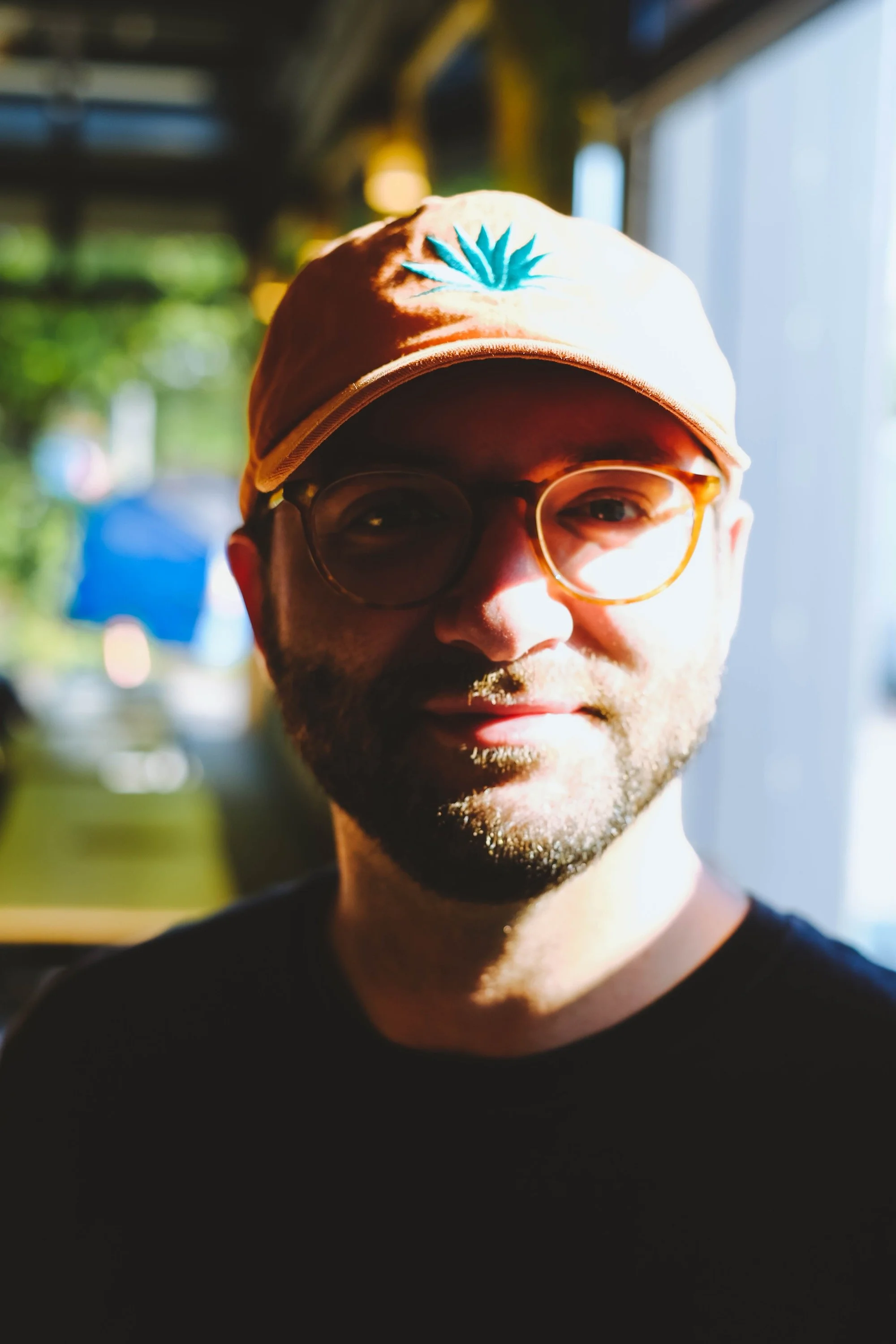 A man with glasses and a baseball cap, sitting in a sunlight-filled space, smiling at the camera.