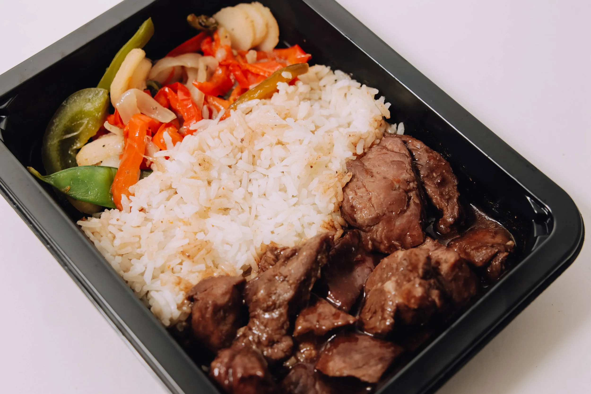 Black lunch container with rice, beef stew, and mixed vegetables including bell peppers, carrots, and cauliflower.