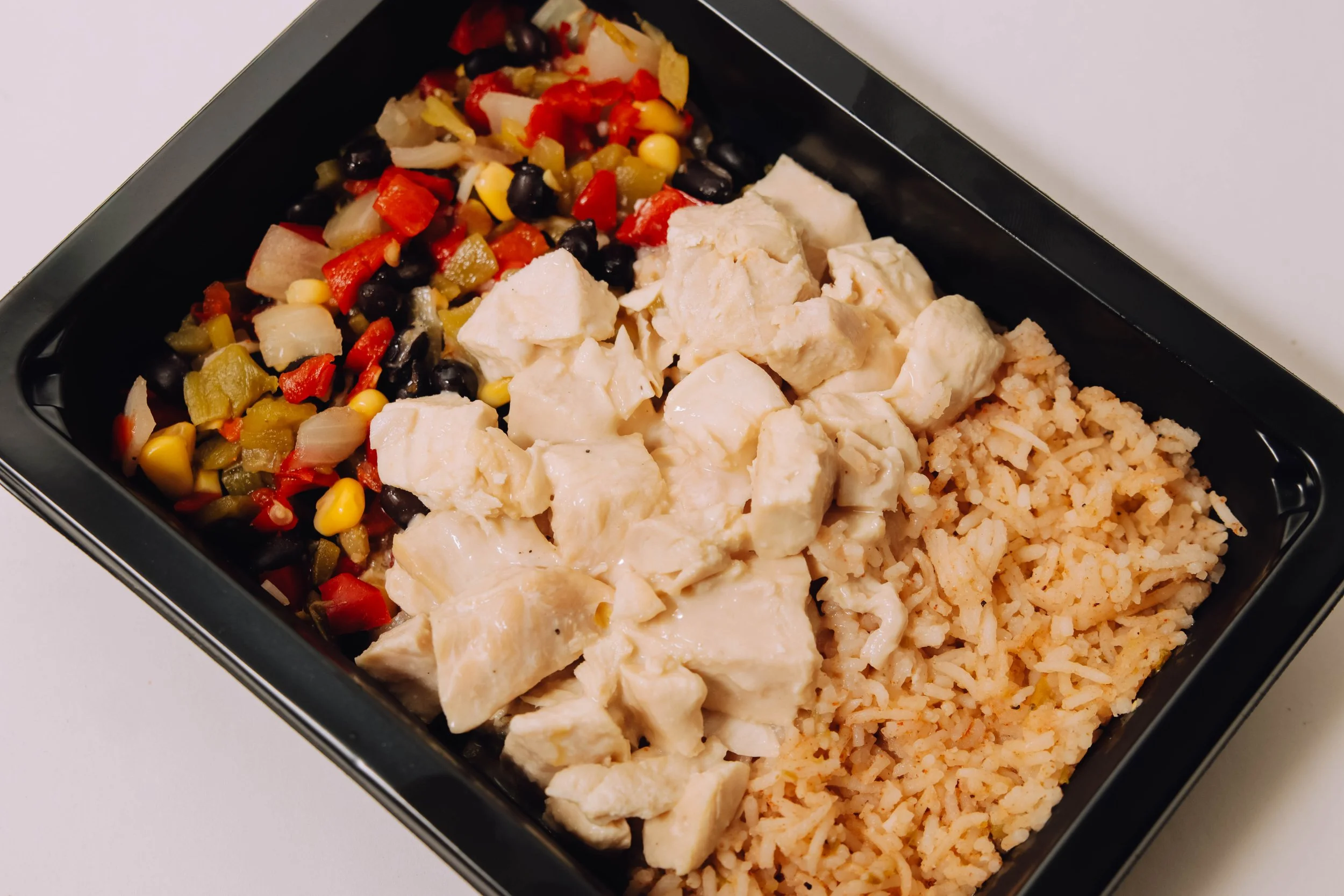 A black meal prep container holding seasoned rice, chicken in white sauce, and a vegetable salad with corn, peppers, onions, and black beans.