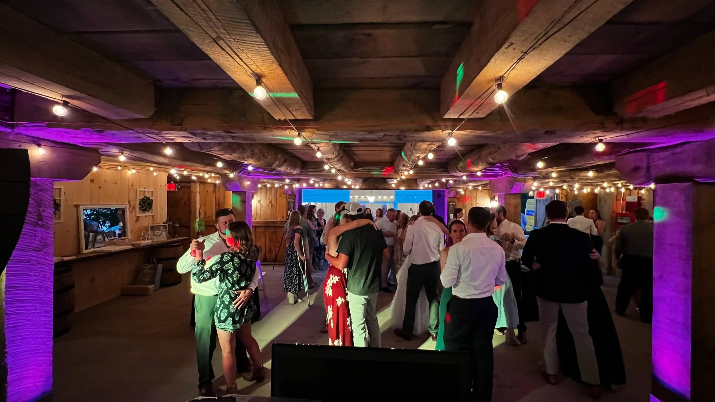 Indoor wedding reception with people dancing and wooden beams, colorful lighting, and string lights.