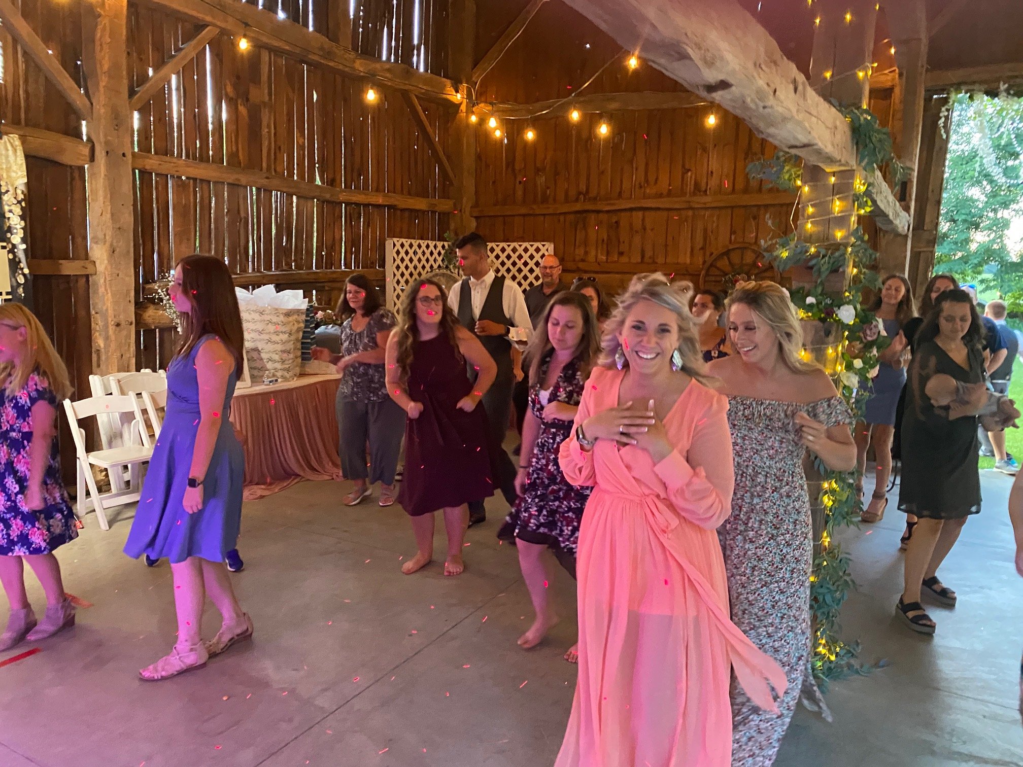 Group of people dancing in a rustic barn with string lights, featuring women in dresses and a lively, festive atmosphere.