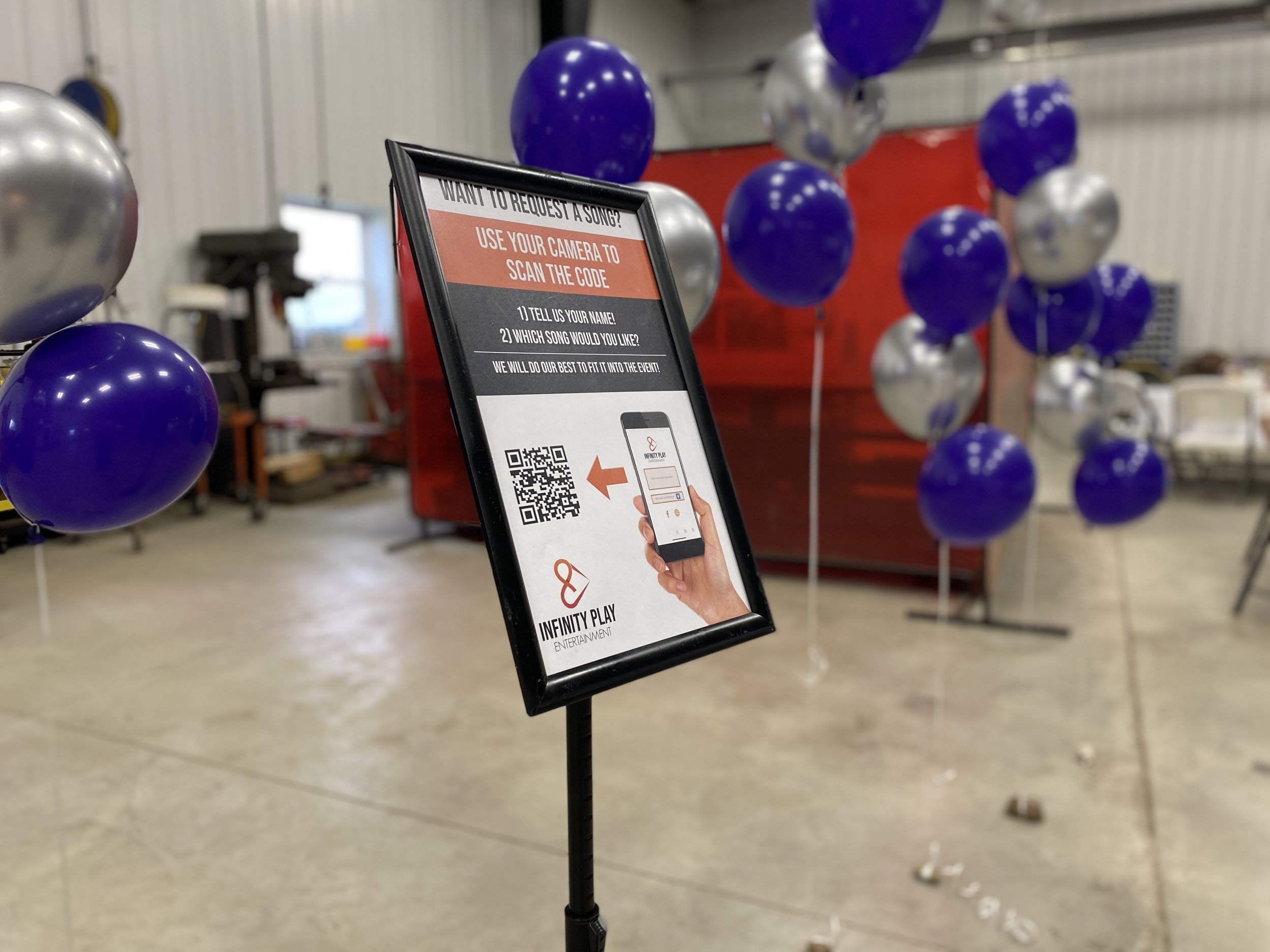 Indoor event setup with purple and silver balloons, a sign for song requests via QR code, and a red backdrop.