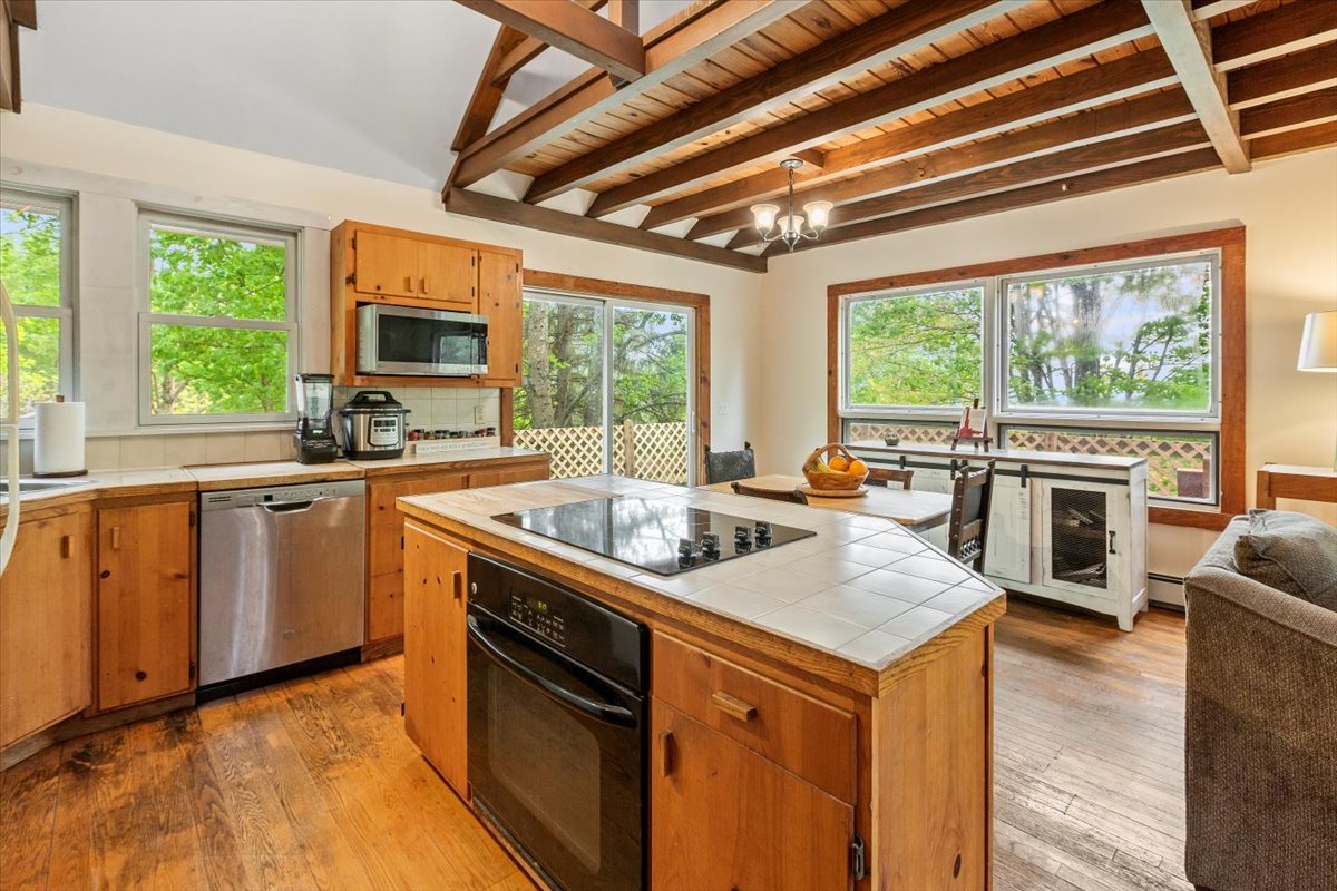 Open kitchen area with island with built-in stove, windows, and wood floors and beam ceiling.