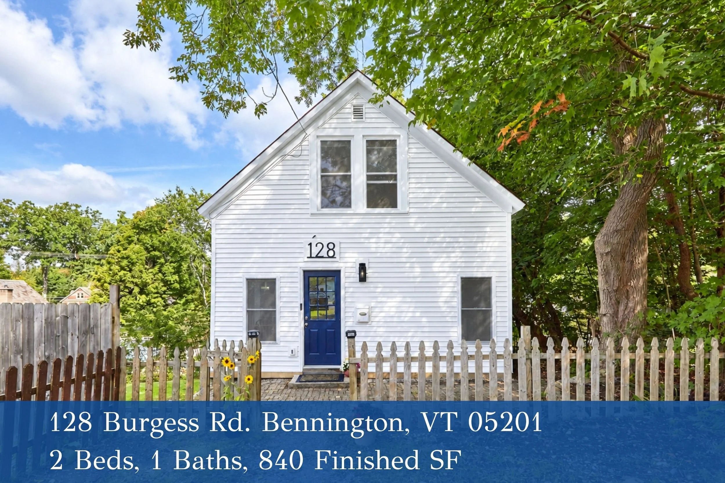 White house with blue door and number 128, surrounded by a wooden picket fence, green trees, partly cloudy sky, and a sign with the address and property details.