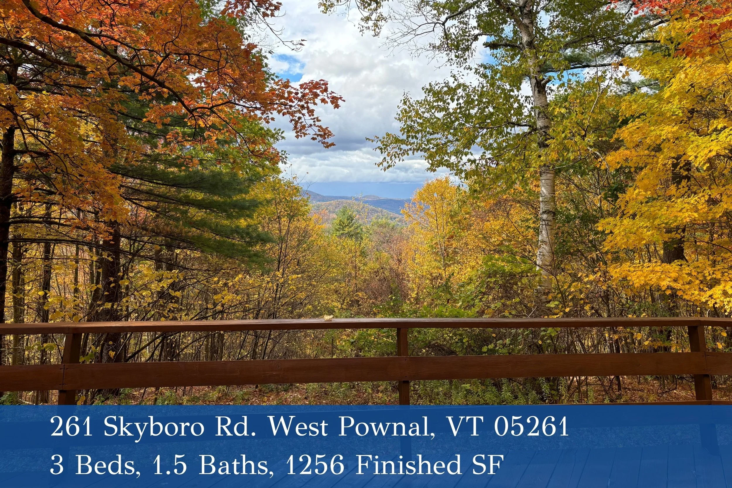 Scenic view of trees with colorful autumn leaves on a cloudy day, overlooking distant mountains and a body of water, from a wooden deck with a railing.