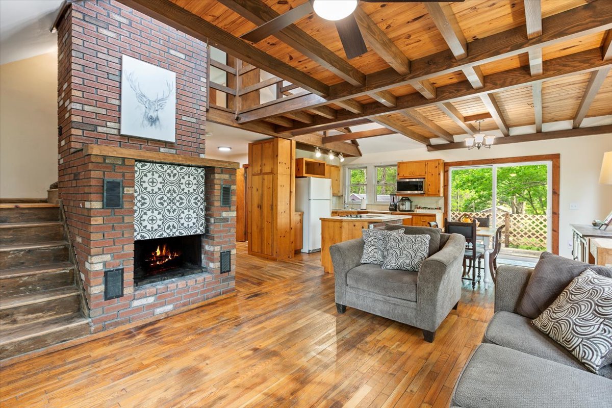 Open living space with fireplace, ceiling fan, wood floors and beamed ceilings.