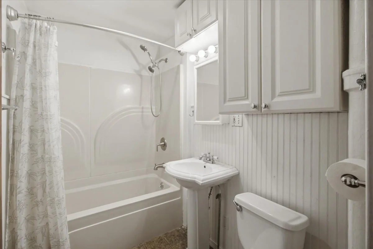Small bathroom with a bathtub and shower, a pedestal sink, a toilet, and cabinetry above the sink, all in white.
