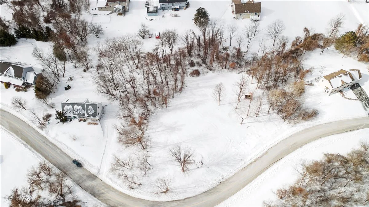 An aerial view of a snowy residential area with houses, trees, and a winding road.