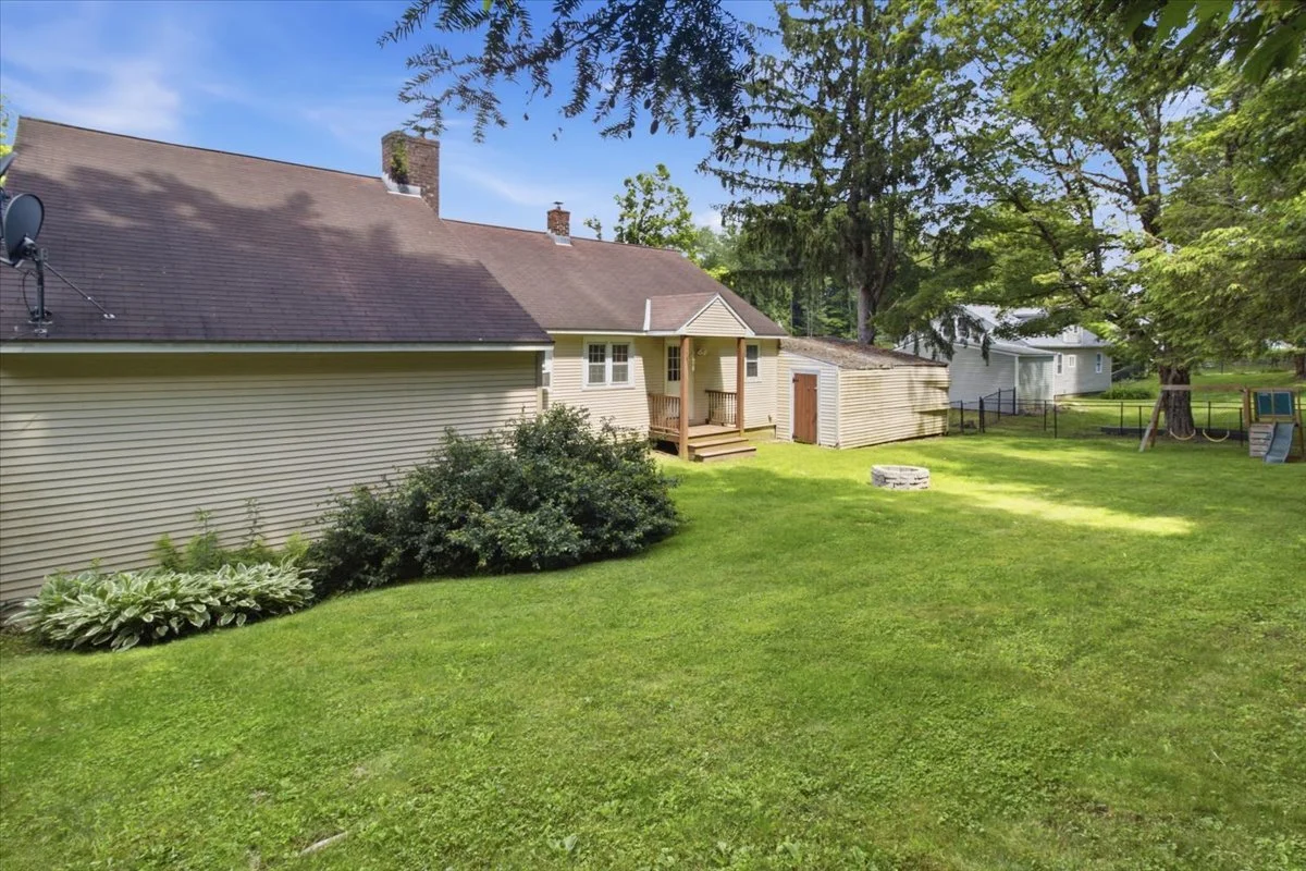 Backyard of a house with green lawn, trees, and a small fire pit. There is a beige house with a small deck and a shed in the yard.