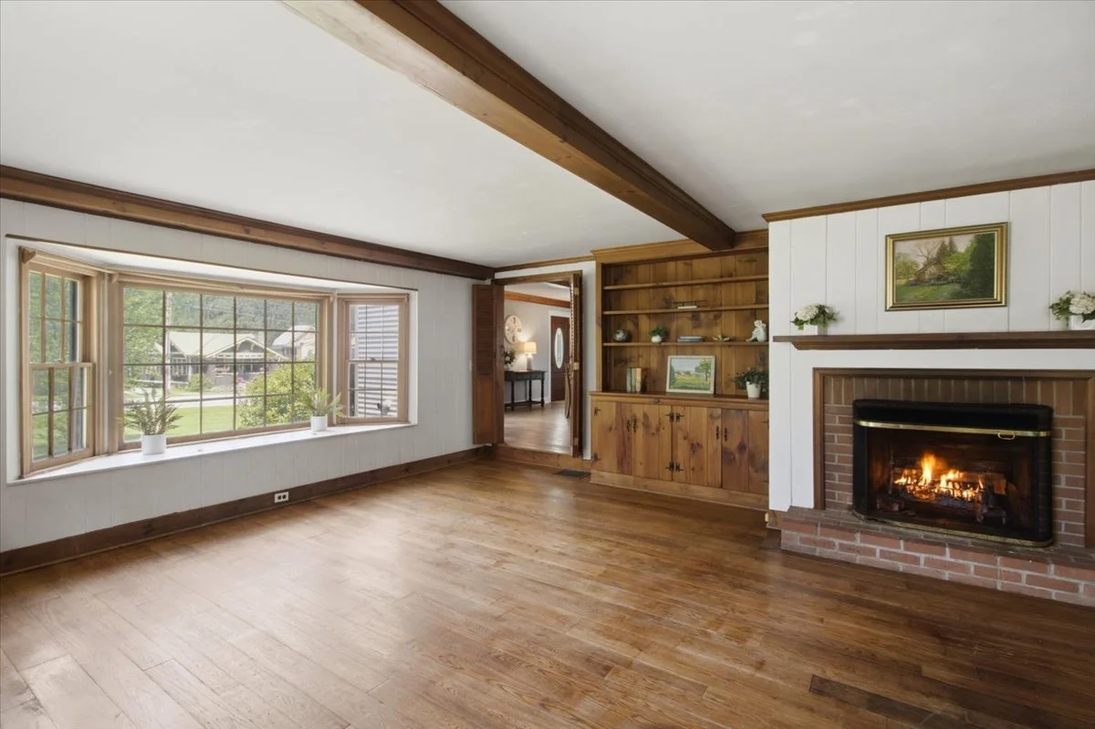 Open living area with fireplace, built ins, exposed cieling beams and large windows allowing in sunlight.