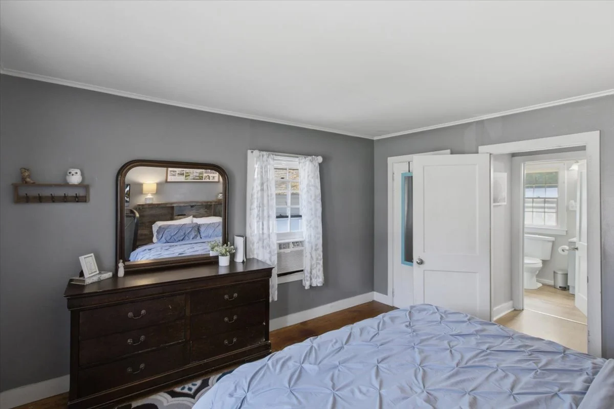 Bedroom with dresser and large mirror, floating shelves. Gray walls, white ceilings and a bed with blue comforter.