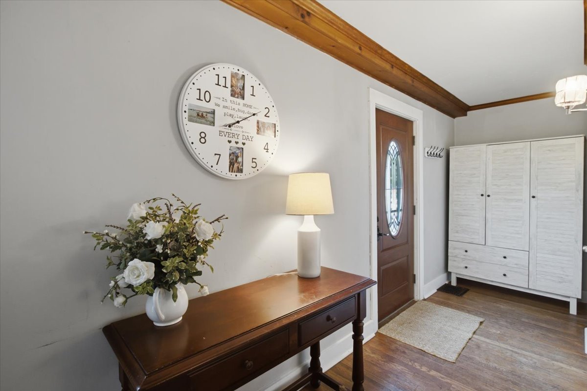 Entryway in Cape Cod style home showing clock, console table with florals and lamp and storage hutch. Distinctive dark floors and wood mouldings visible near front door with decorative oval window.