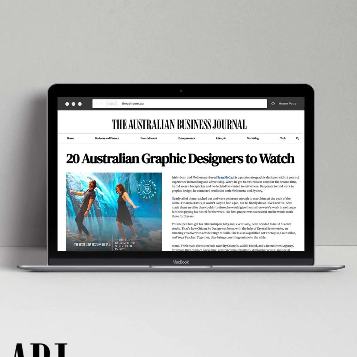 Laptop displaying an article from The Australian Business Journal titled '20 Australian Graphic Designers to Watch', featuring a photo of two people posing against a blue and teal background.