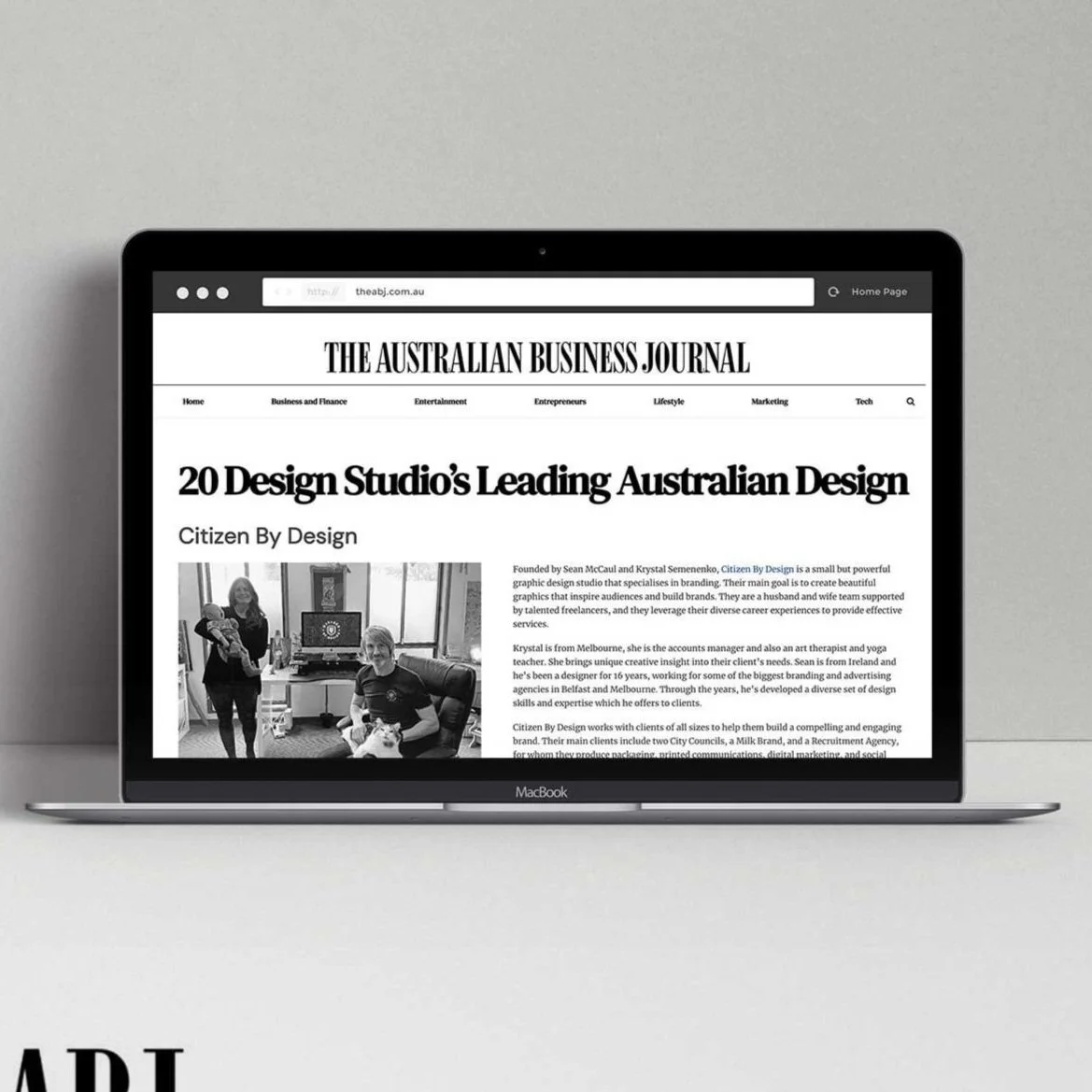 Laptop displaying a news article from The Australian Business Journal about a design studio, with a black and white photograph of two people in a creative workspace.