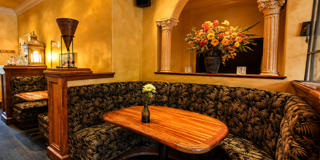 A cozy restaurant corner with a curved wooden booth, a small wooden table with a single white flower in a dark vase, a large floral arrangement in a dark vase on the ledge behind the booth, warm yellow walls, decorative columns, and soft lighting.