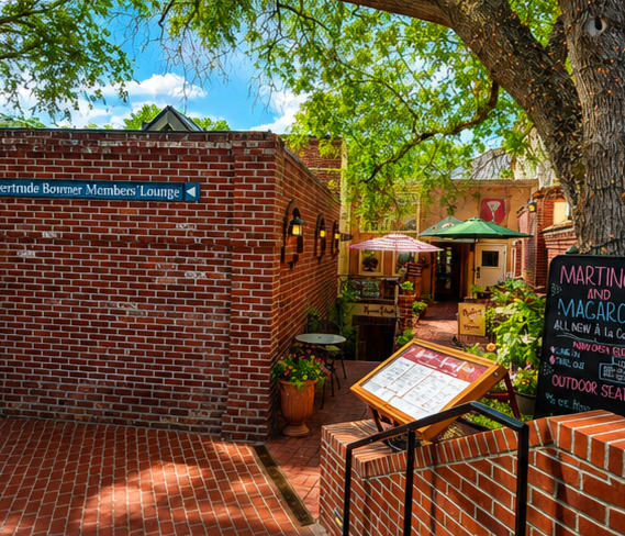 Outdoor patio area of a restaurant or cafe with tables, umbrellas, plants, and a brick wall. A sign points toward the Bower Members Lounge. A chalkboard menu is visible.