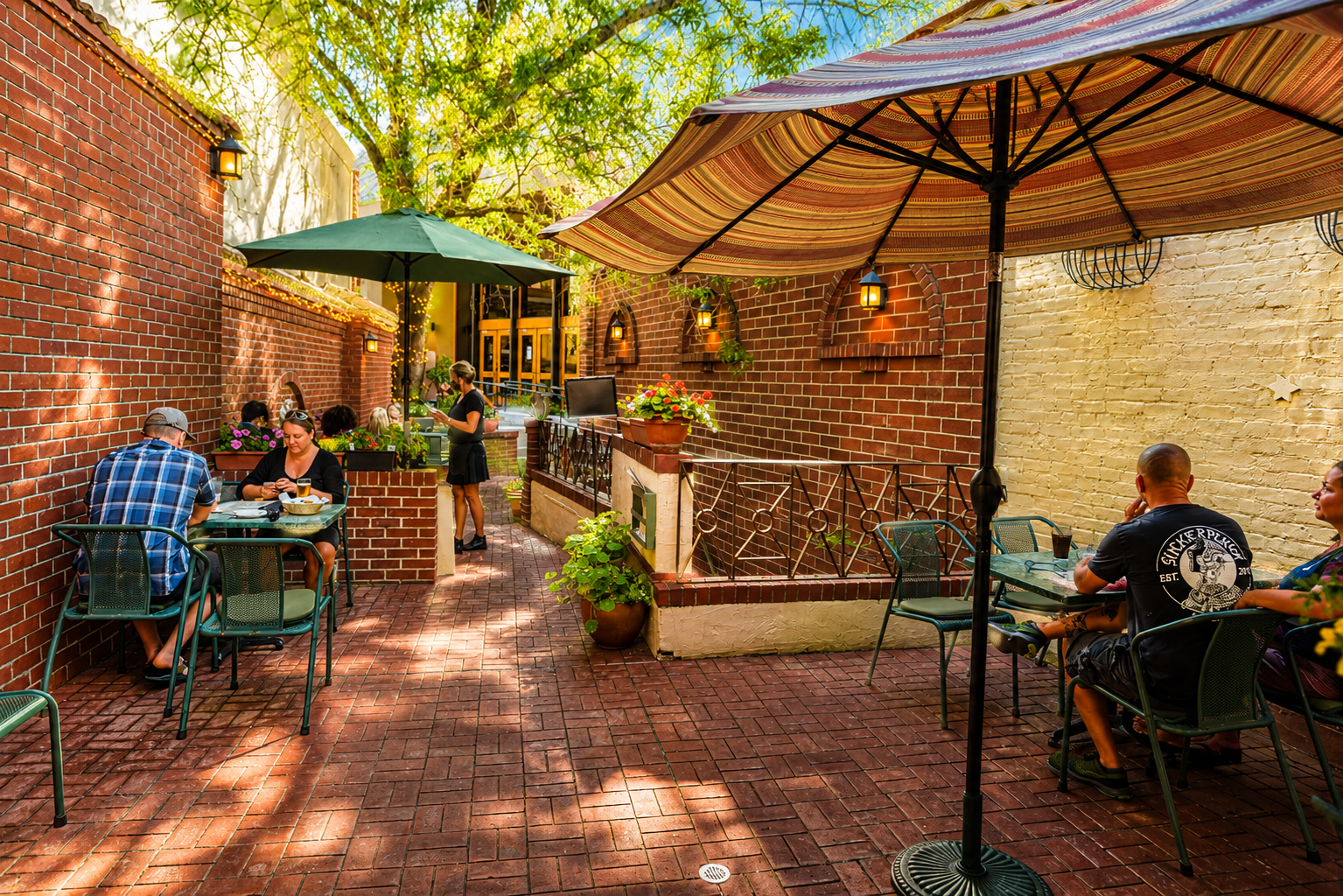 An outdoor patio of a restaurant with brick walls, multiple tables and chairs, large umbrellas providing shade, and people dining and talking.
