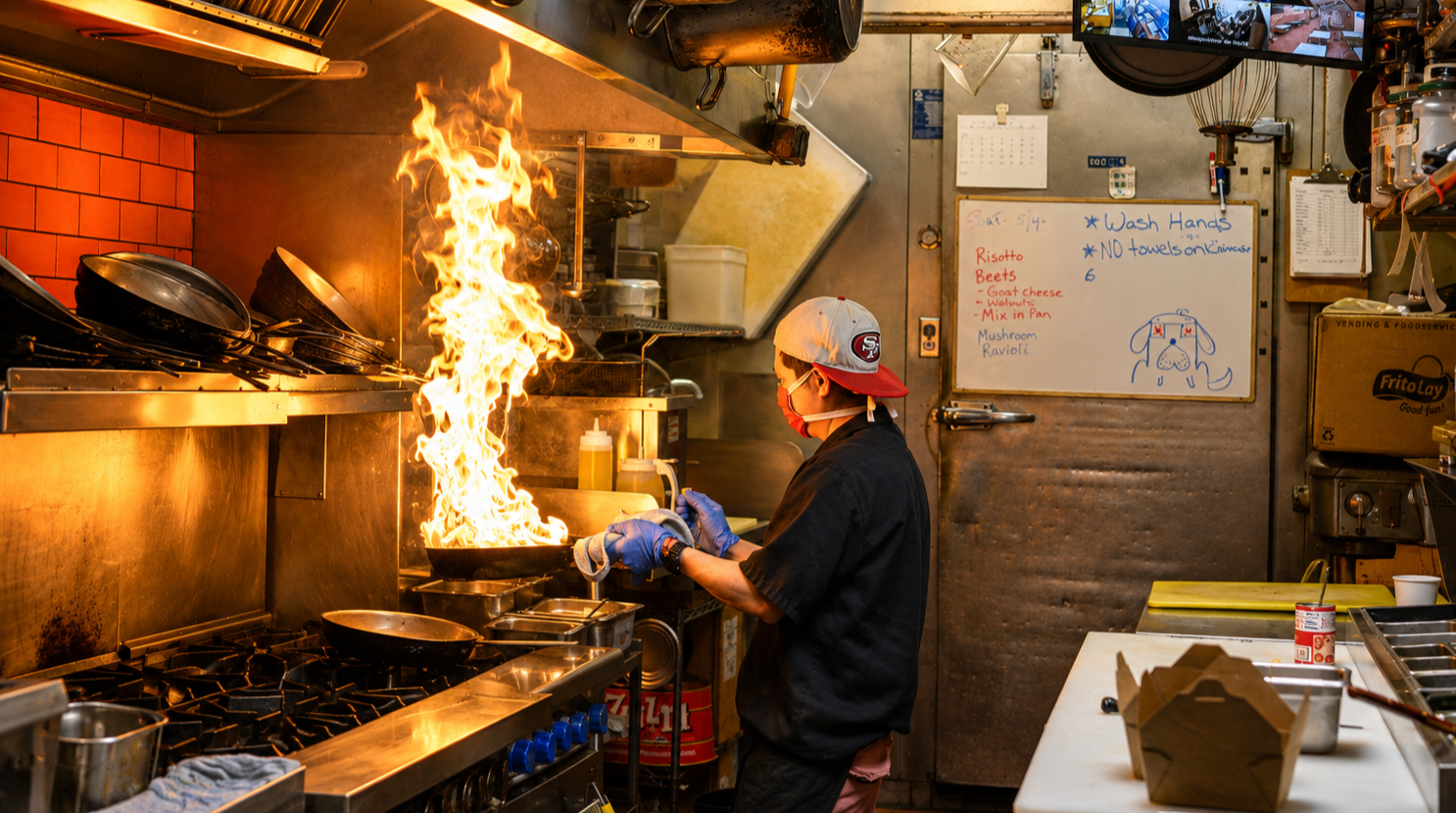 A chef cooking in a busy restaurant kitchen with a large flame rising from the stove, wearing gloves, a face mask, and a baseball cap.