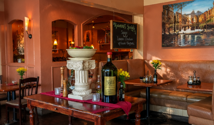 Interior of a cozy restaurant with leather booths, tables decorated with yellow flowers, a large wine bottle on a wooden table, and a chalkboard menu.