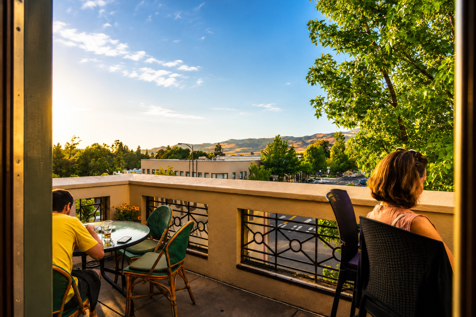 People sitting on a balcony during sunset, looking at the view of trees, buildings, streets, and distant hills under a partly cloudy sky.