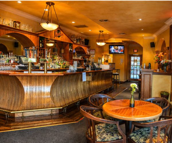 Interior view of a cozy bar with a wooden curved counter, hanging pendant lights, a small round table with four chairs, and a television mounted on the wall, decorated with flowers and warm colors.