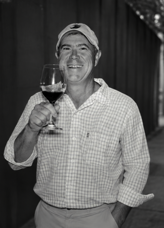 A man smiling and holding a glass of red wine, wearing a baseball cap and a checkered button-up shirt, standing outdoors.