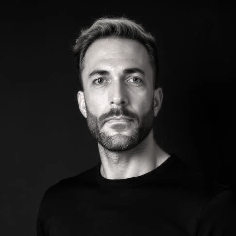 Black and white portrait of a man with short hair and beard, wearing a black shirt, looking at the camera against a dark background.